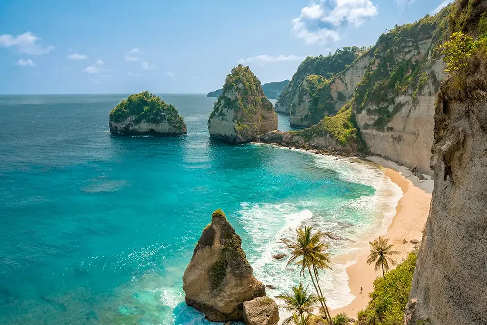 Tropical beach with clear blue water, large rock formations, and green cliffs along the shoreline, with palm trees in the foreground.