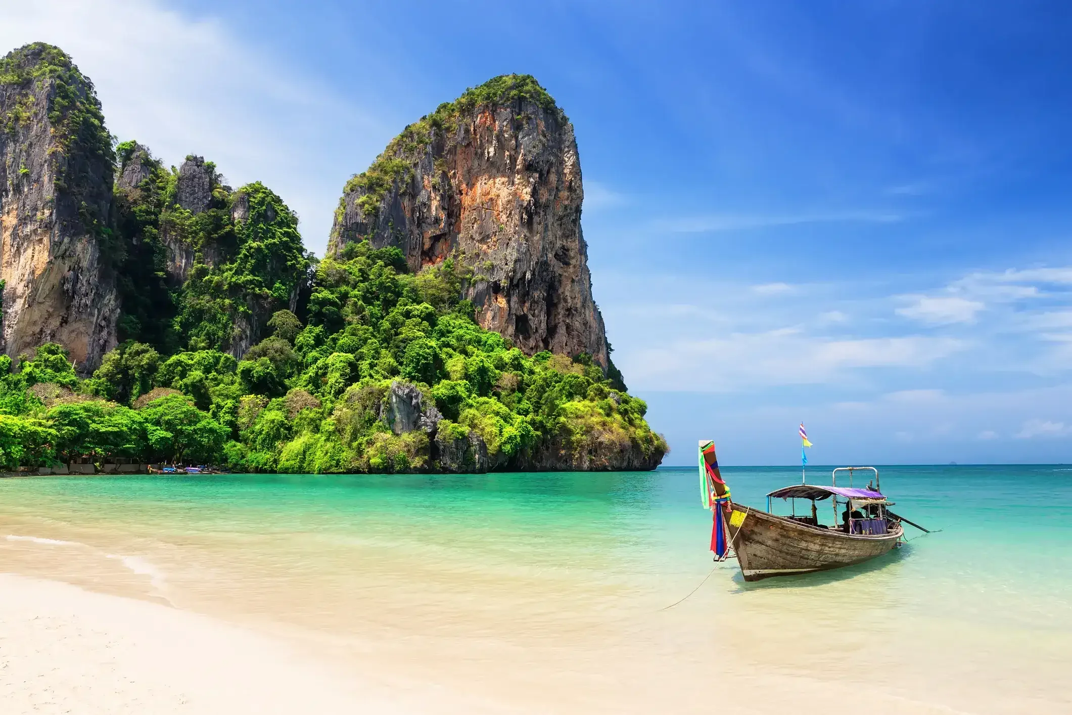A tropical beach with turquoise water, white sand, a longtail boat with colorful decorations, lush green cliffs, and clear blue sky.