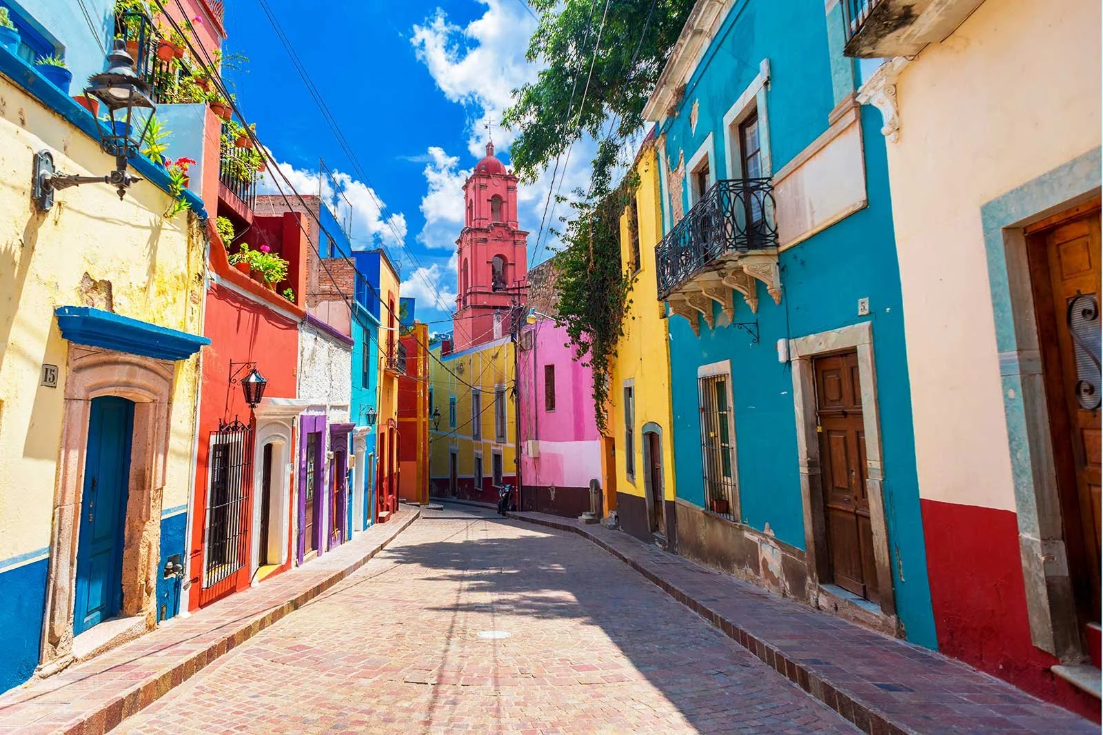 Colorful buildings lining a narrow cobblestone street in a vibrant neighborhood, with a pink church tower in the background, under a partly cloudy blue sky.