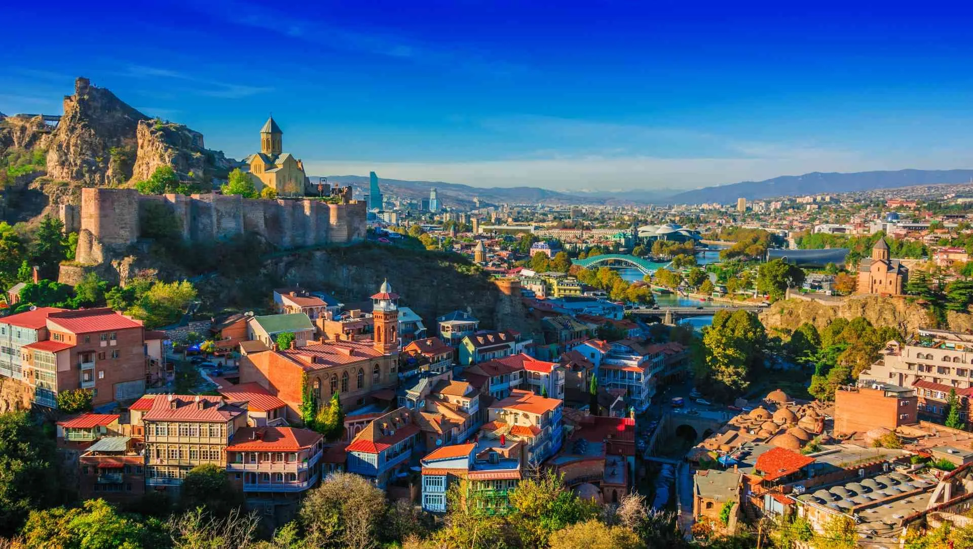 A panoramic view of a city with colorful buildings, a river, historic structures, and hills in the background under a bright blue sky.