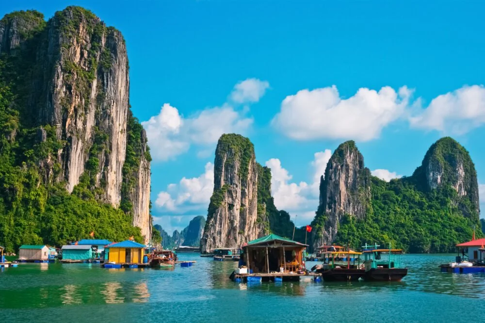 Scenic view of a bay with turquoise water, surrounded by towering limestone cliffs covered in green vegetation. Several floating houses and boats are visible in the foreground, with a partly cloudy blue sky overhead.