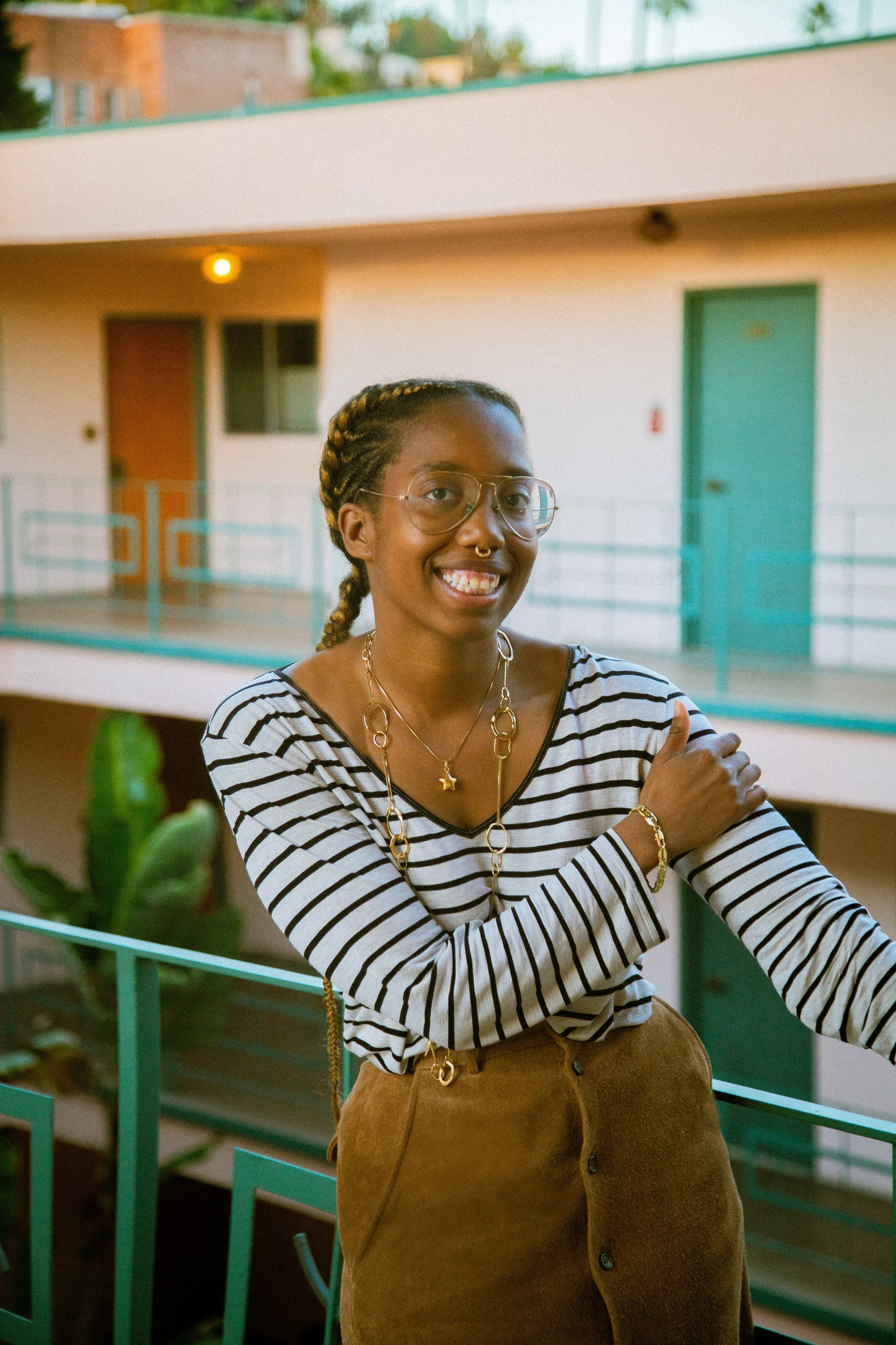 Smiling woman with braids, glasses, and layered gold jewelry standing outdoors on a balcony, wearing a striped shirt and brown pants, with apartment buildings in the background.