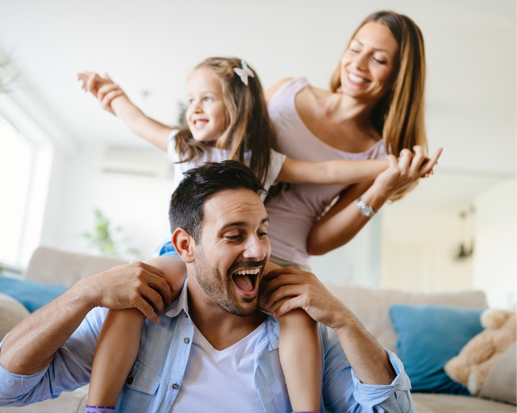 A father giving his daughter a piggyback ride at home, mother smiling alongside, all of them laughing.