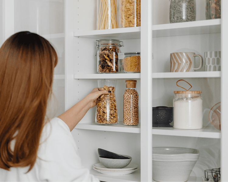 A woman organizing a bright white pantry with neatly arranged glass jars of grains, nuts, and pasta.