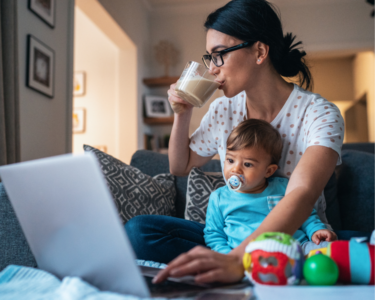 A mother sipping coffee with her baby in her lap and a laptop open in front of her.