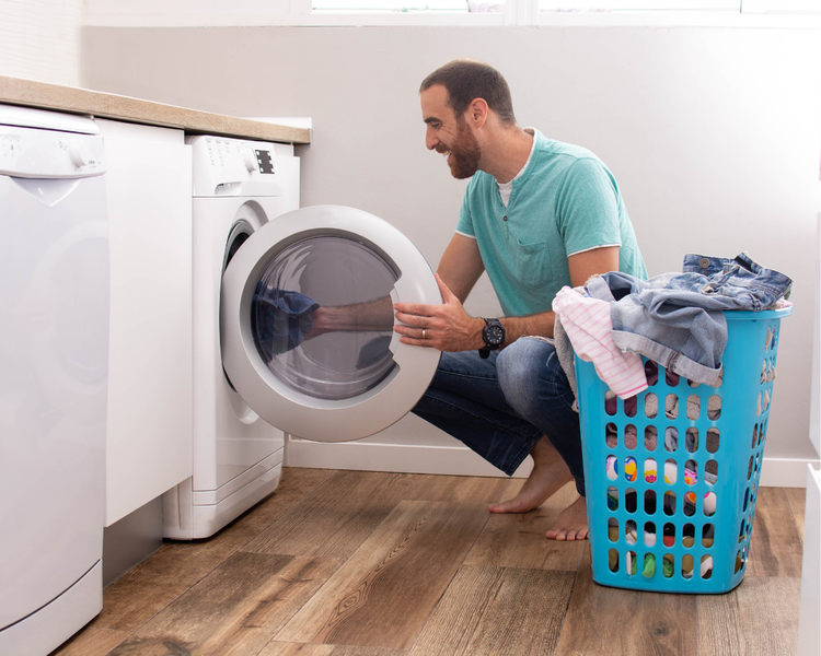 A man kneeling at a front-loading washing machine, loading laundry from a basket of clothes.