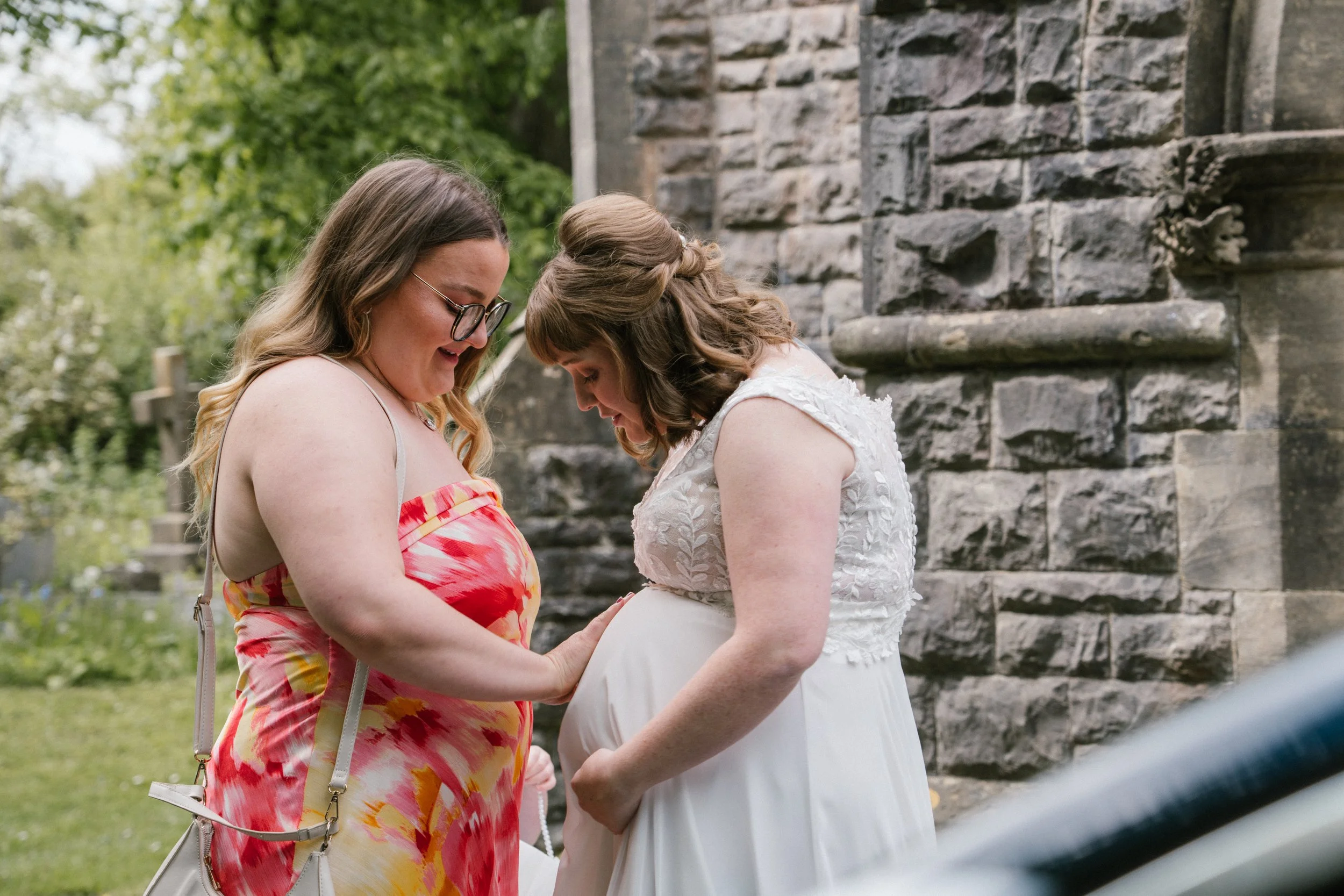 Two women share a tender moment outdoors, one appearing to be pregnant, with one woman placing her hand on the other's belly. They are smiling and looking at each other. The scene is set beside a stone wall and surrounded by greenery.