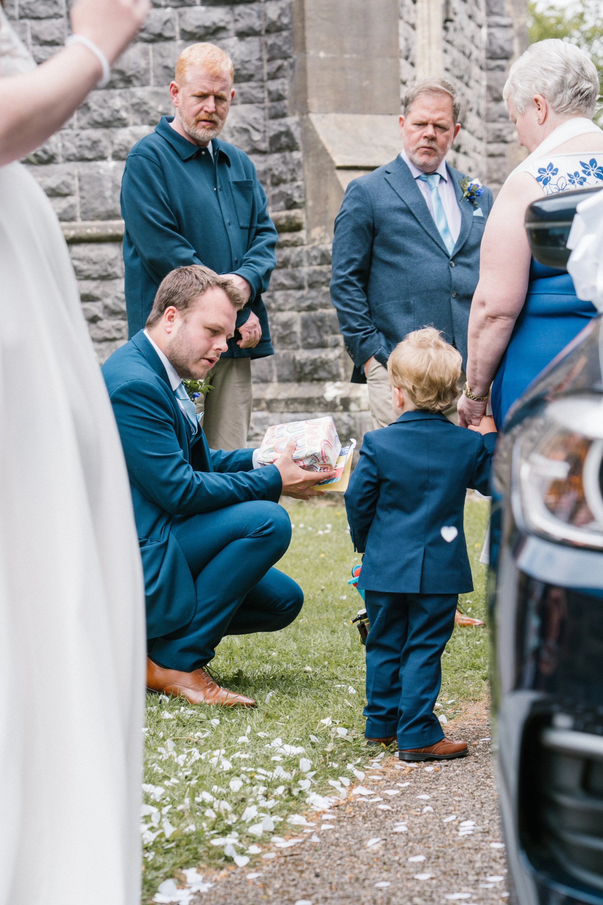 A man in a blue suit kneeling and handing a wrapped gift to a young boy in a blue suit, at an wedding event with guests, stone building, and cars nearby.
