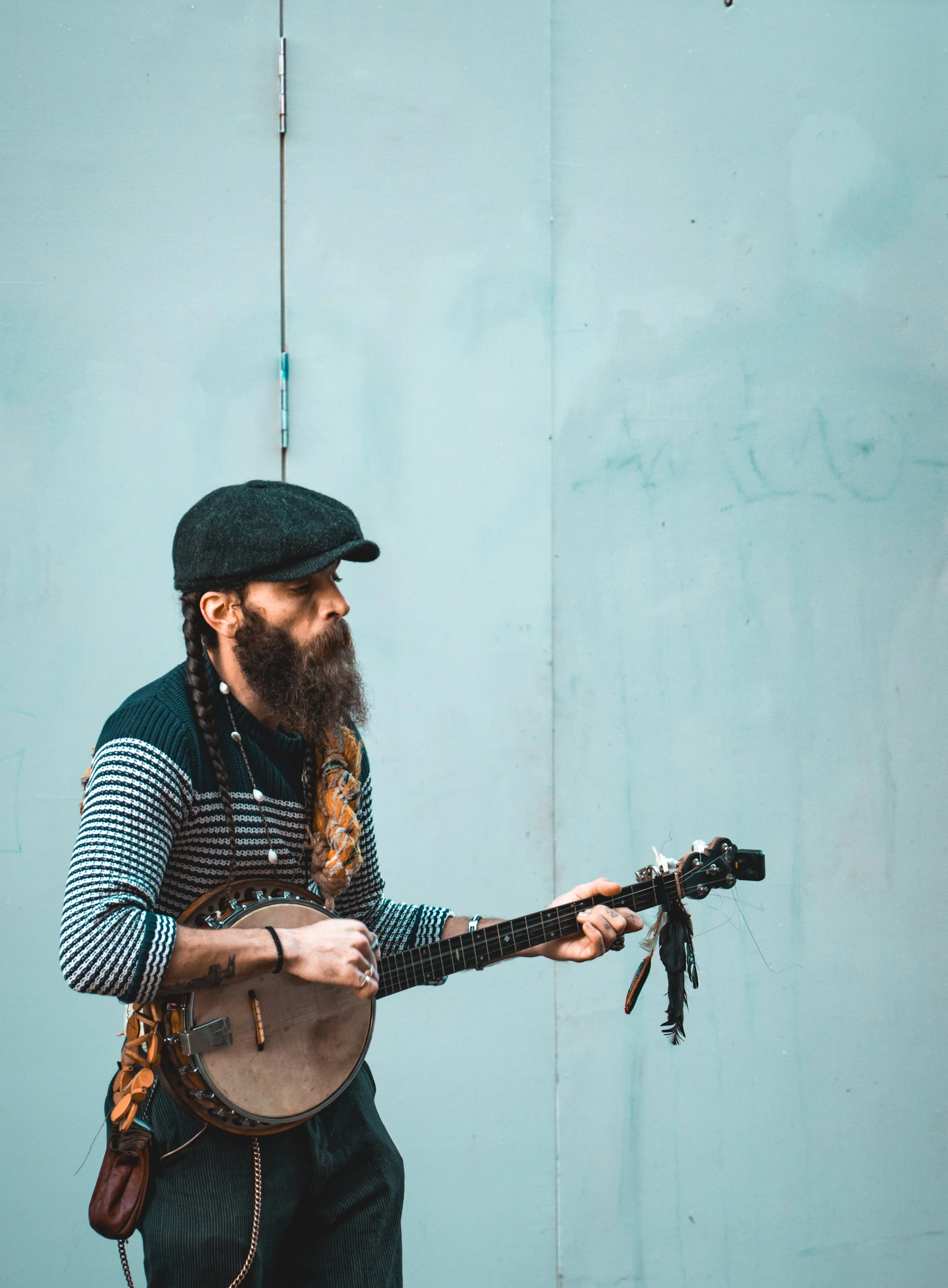 A bearded man wearing a dark gray cap, striped sweater, and dark pants playing a guitar and a banjo against a plain light-colored wall.