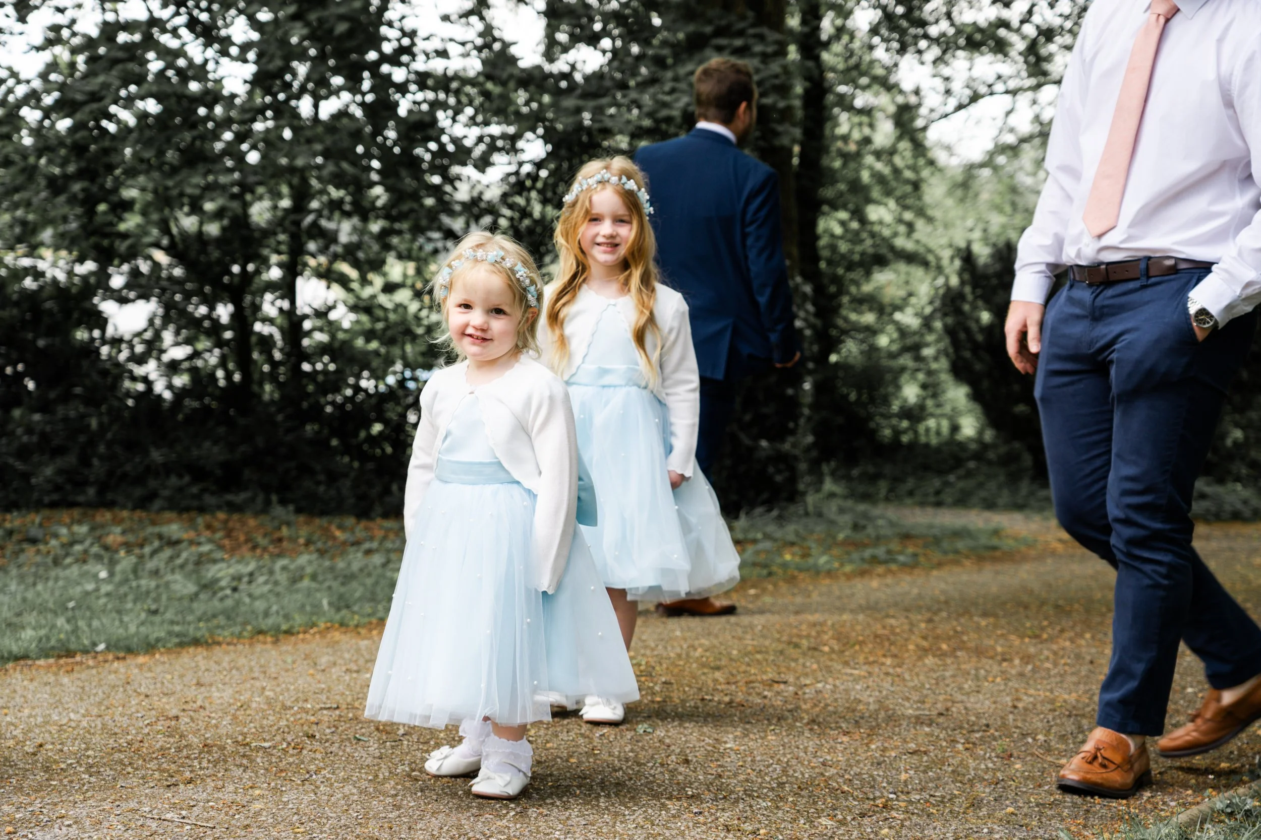 Two young girls in white dresses with floral headbands standing outdoors, with adults nearby, flower girls for a wedding