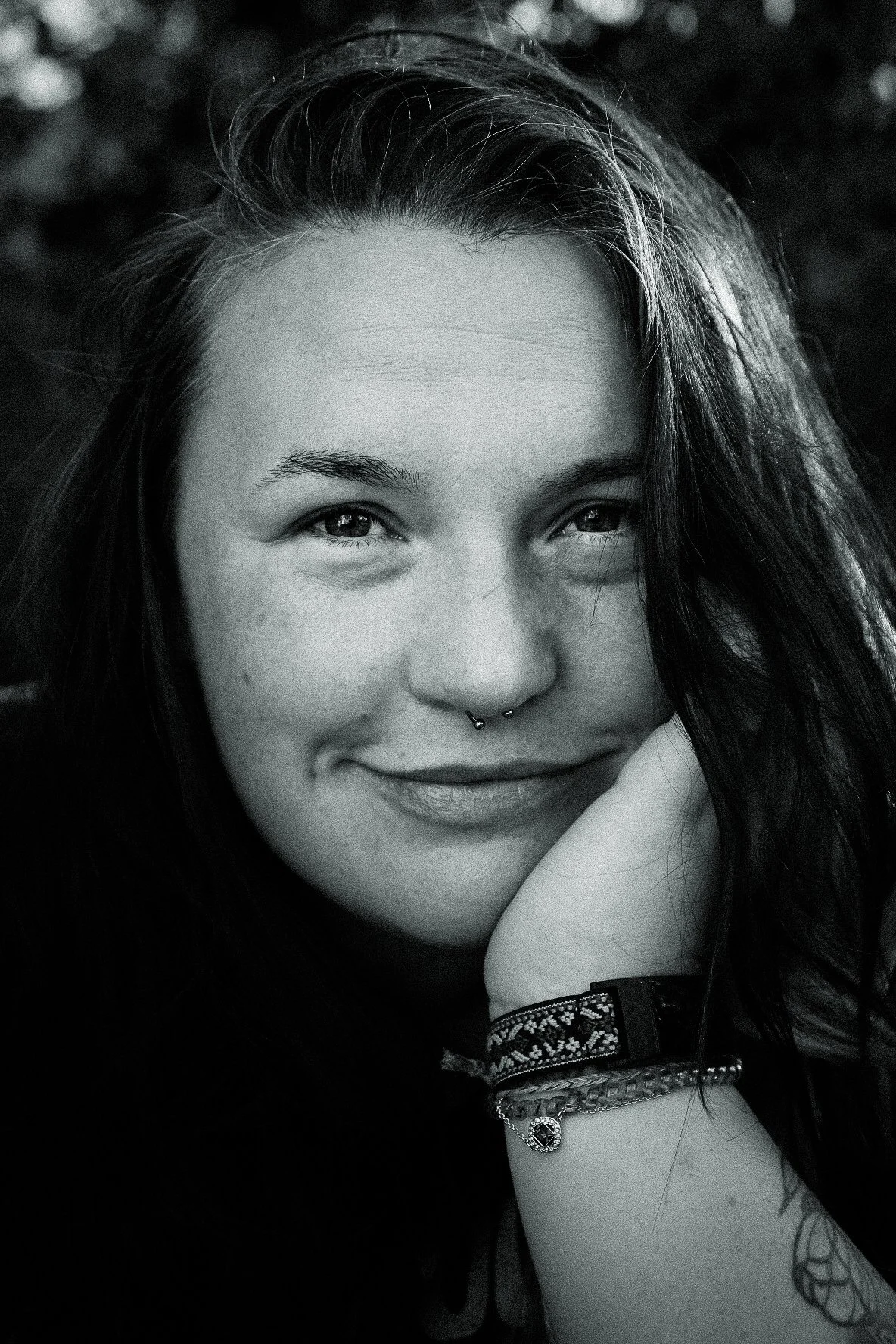 Close-up black and white portrait of a young woman with long, dark hair, smiling gently, wearing a septum piercing, a decorative bracelet, and a necklace.