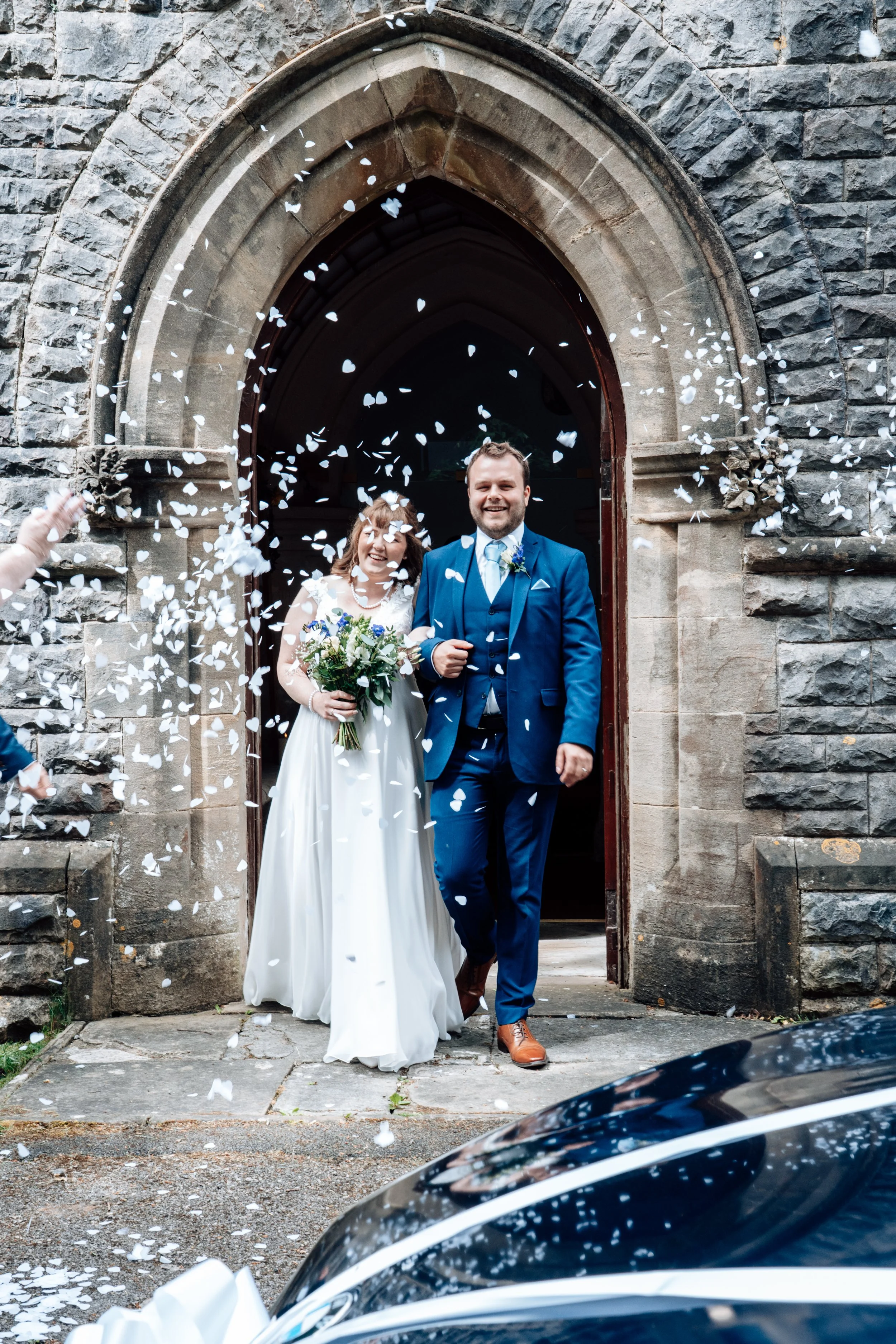 A newlywed couple exiting a stone church while confetti is falling around them. The bride is holding a bouquet and wearing a white wedding dress, and the groom is dressed in a blue suit. They are smiling happily.
