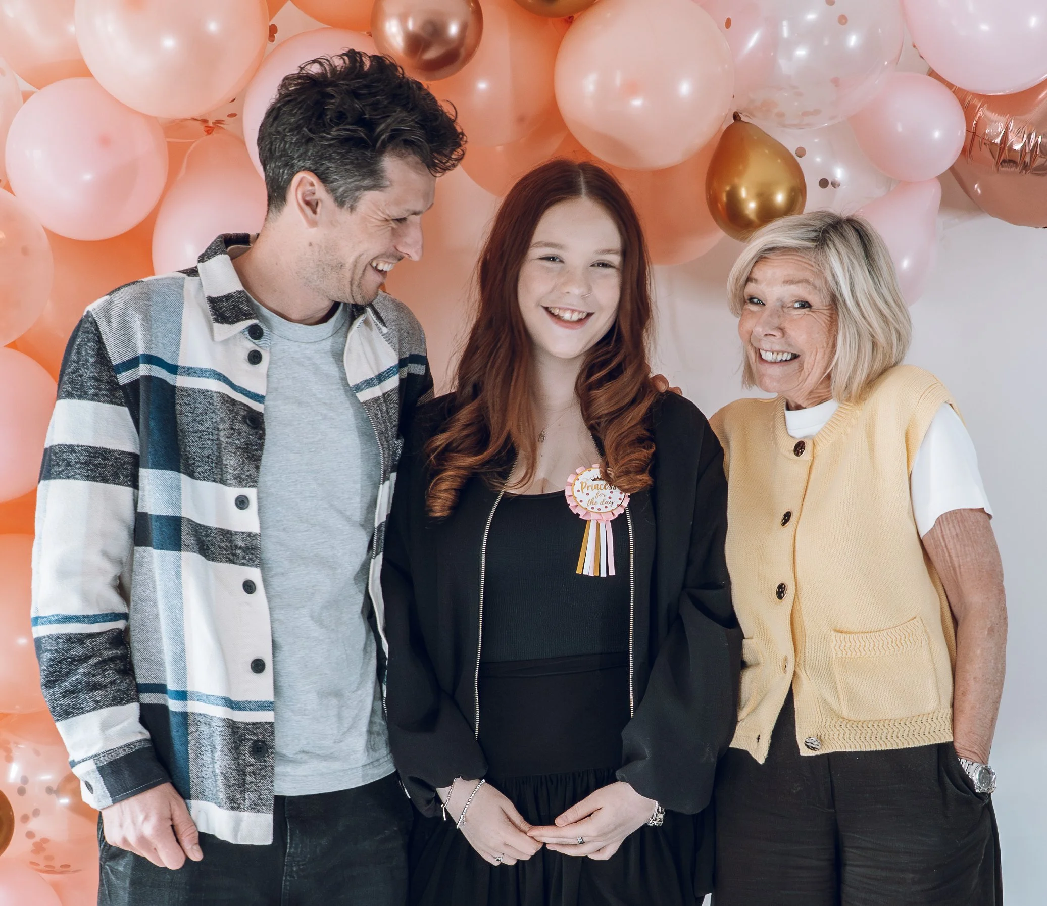 Two older family members smile comfortably with a younger birthday girl. They are standing under a balloon arch and the colour palette is attractive