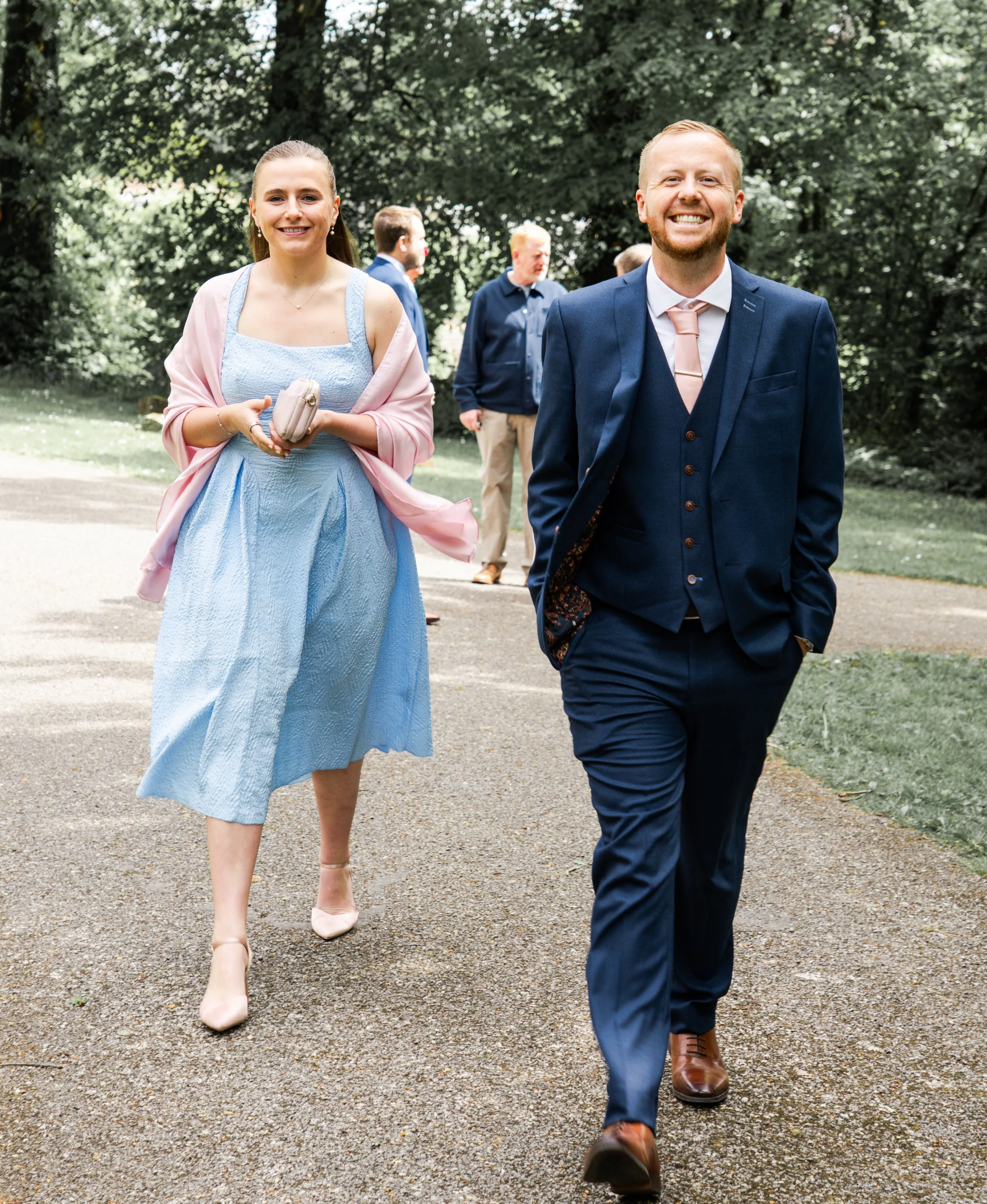A man and woman dressed in formal attire walking outdoors on a sunny day, with other people in the background.