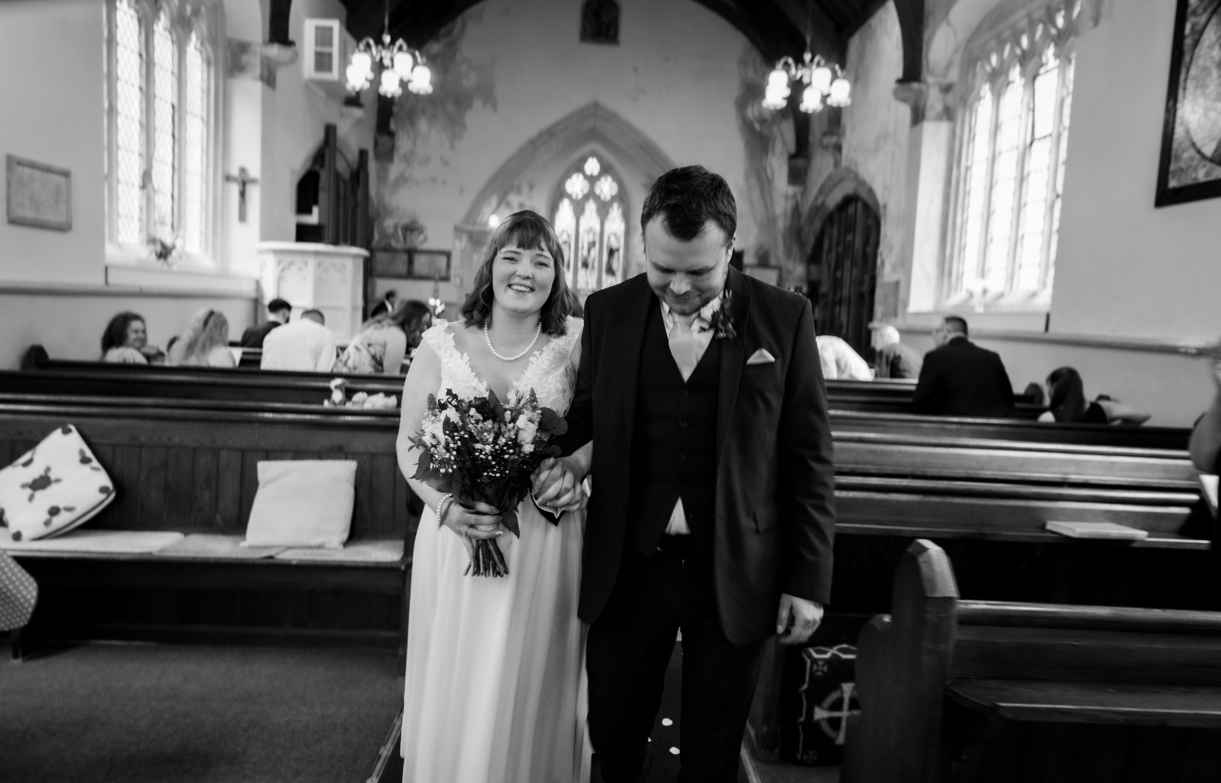 A bride and groom walking down the aisle in a church during their wedding ceremony. The bride is smiling and holding a bouquet, while the groom looks down. They are dressed in wedding attire, and there are guests seated on pews in the background.