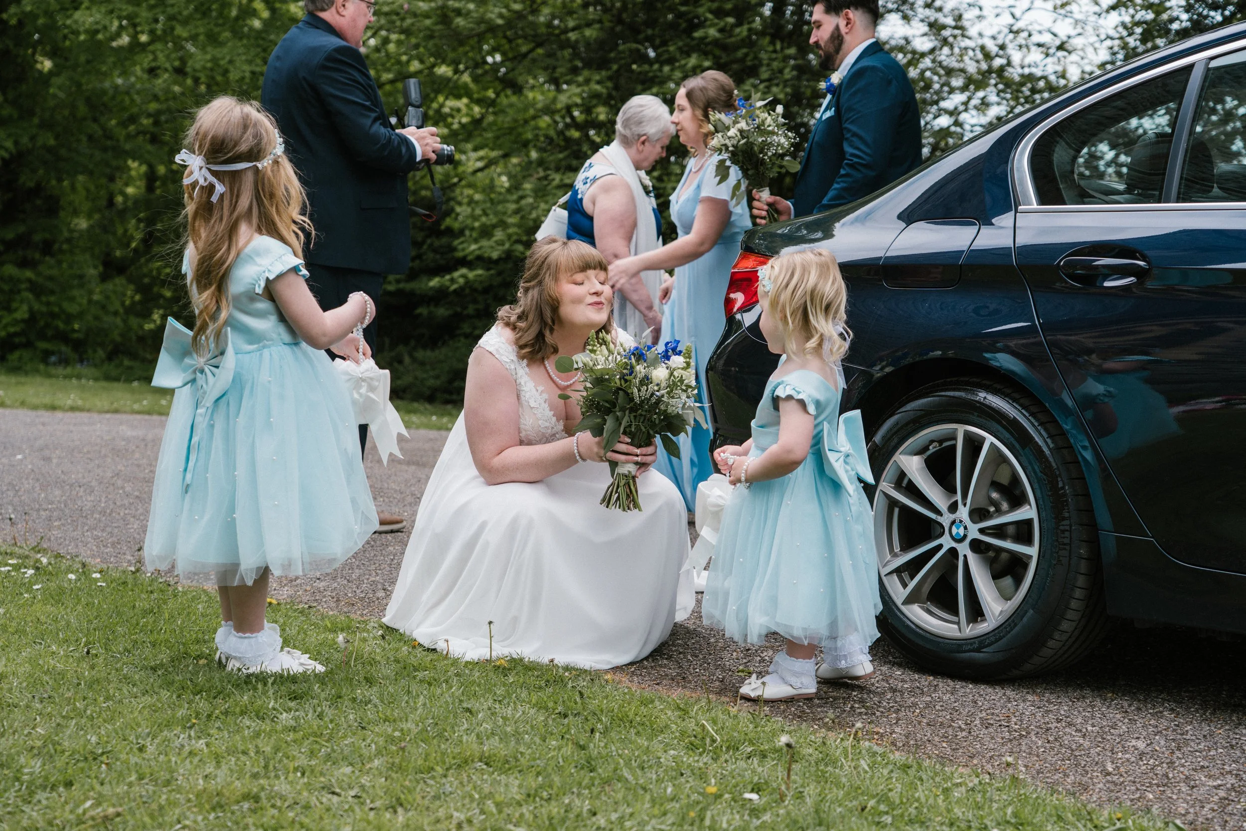 A wedding scene outdoors with a woman in a white dress holding a bouquet, kneeling near a black BMW car next to two young girls in light blue dresses, and a bride and groom in blue attire standing nearby. An older woman and a man with a camera are al