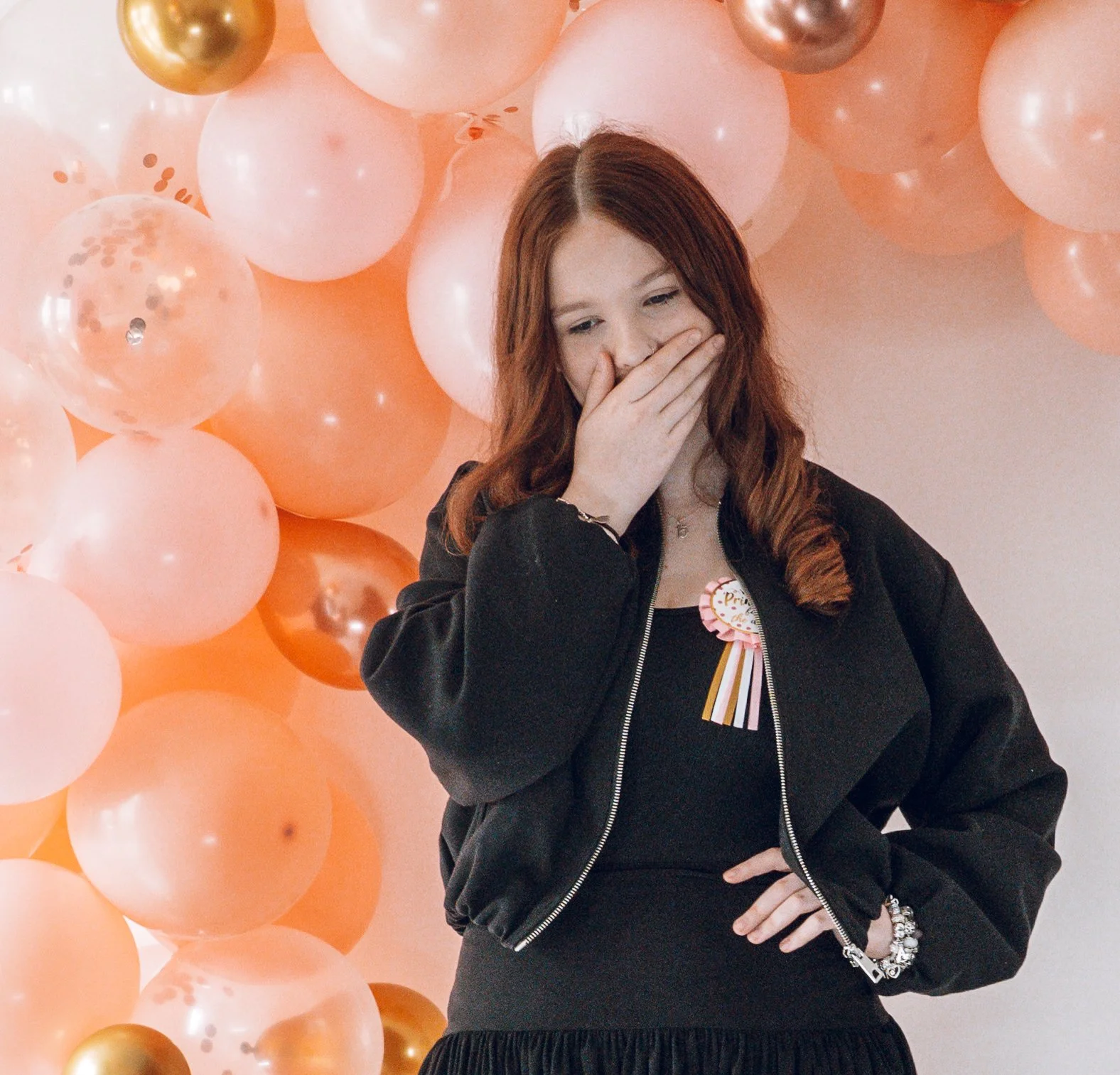 A teenage girl covering her mouth, her emotion is not completely conveyed in this photo. The colour palette is orange and black and rose gold