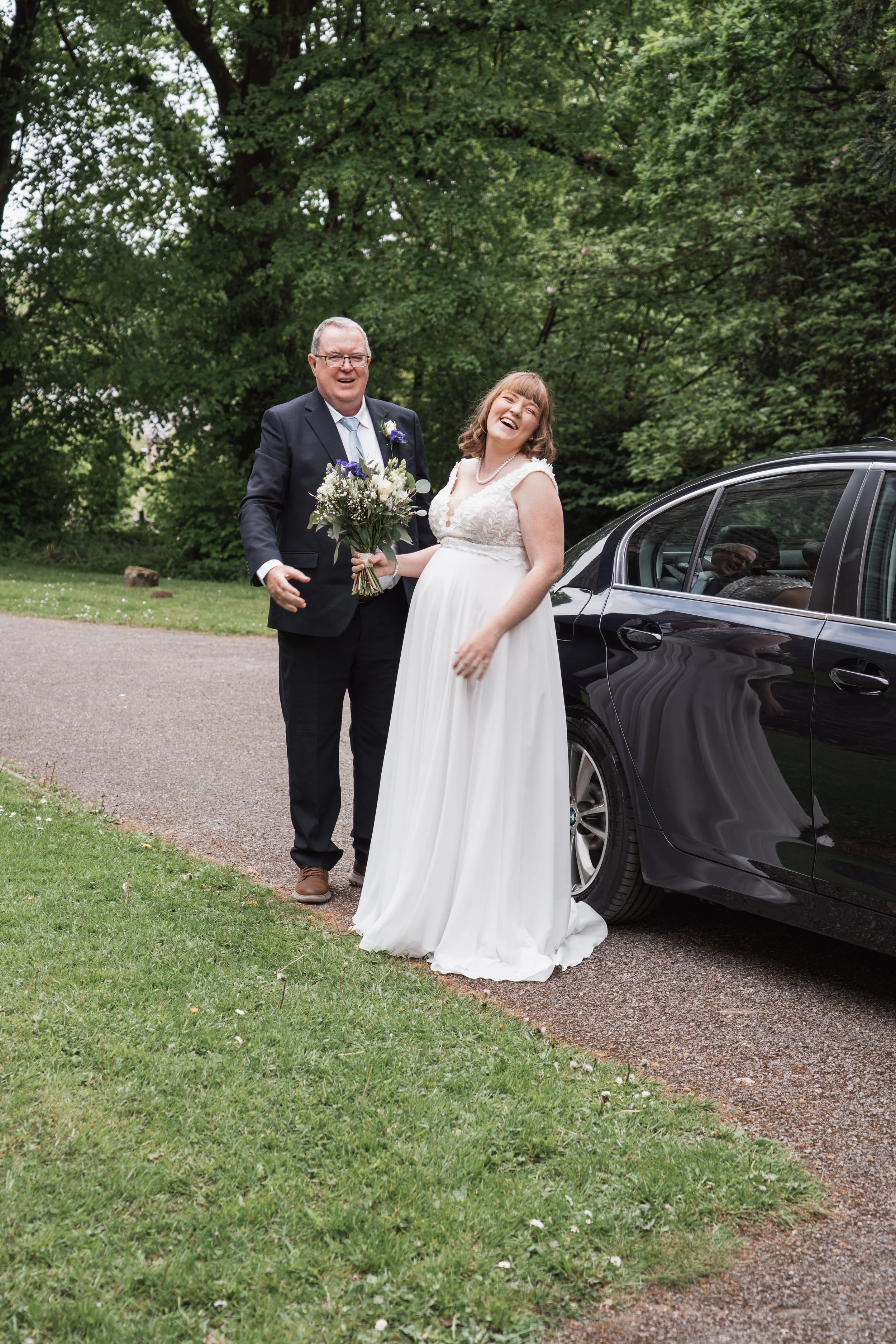A bride and an older man, possibly her father, standing outdoors near a black car. The bride is wearing a white wedding dress and holding a bouquet of flowers, smiling and laughing. The older man is dressed in a dark suit, white shirt, and tie, smili