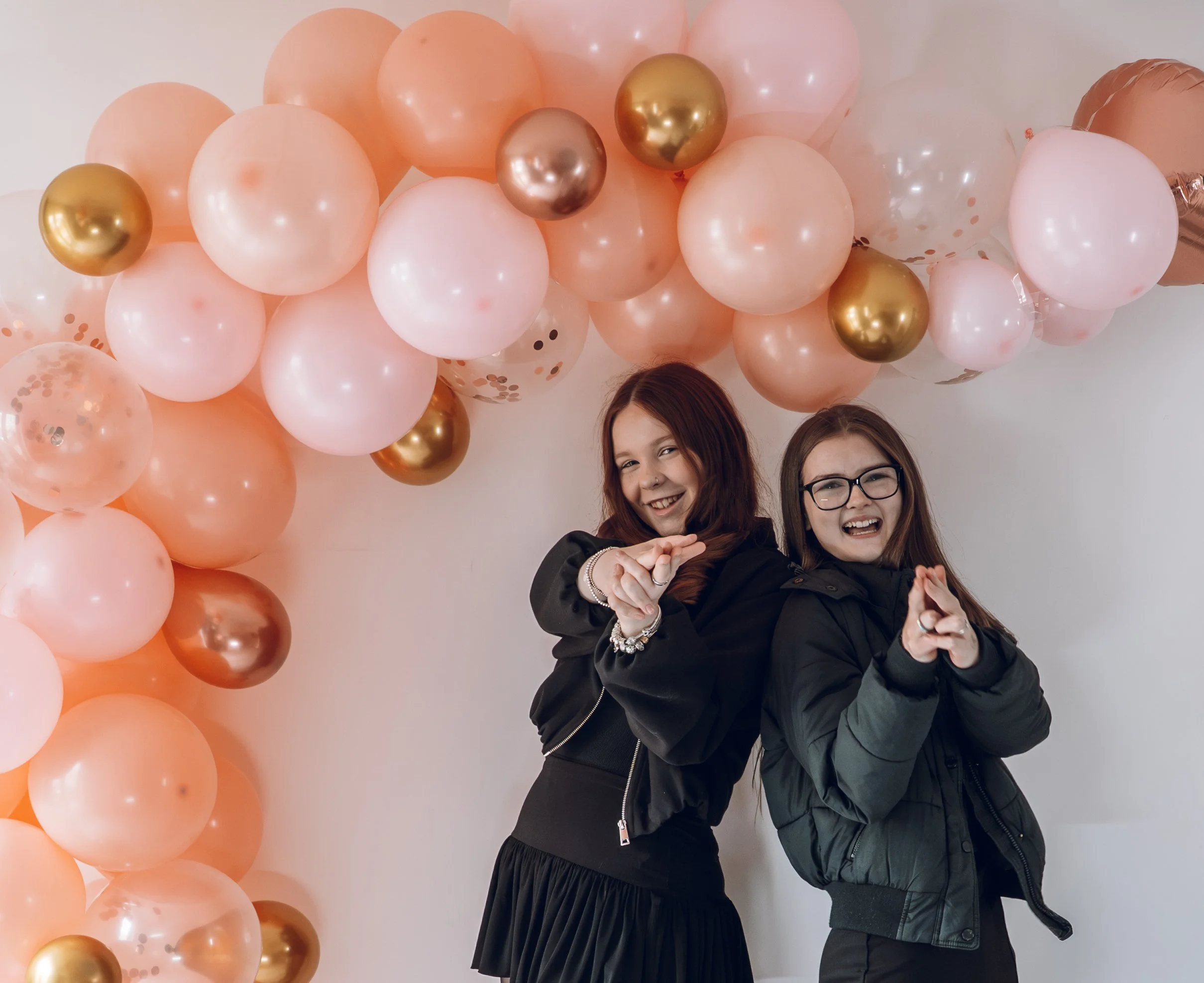 Two young girls standing beneath a balloon arch sending finger guns at the camera. Both look happy and relaxed