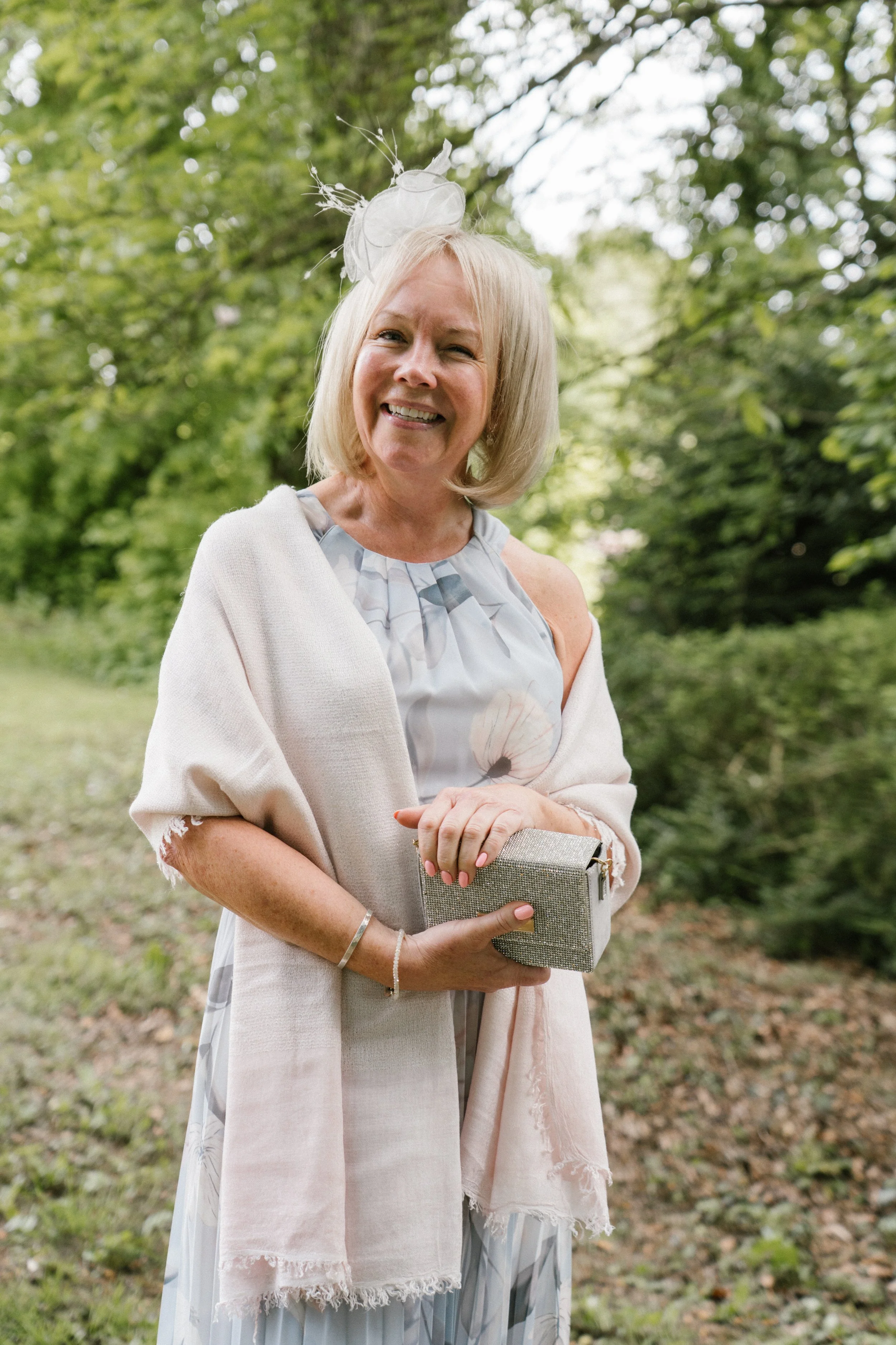 A smiling older woman outdoors, dressed in elegant pastel attire with a fascinator hat, holding a clutch purse, standing in a lush green park.