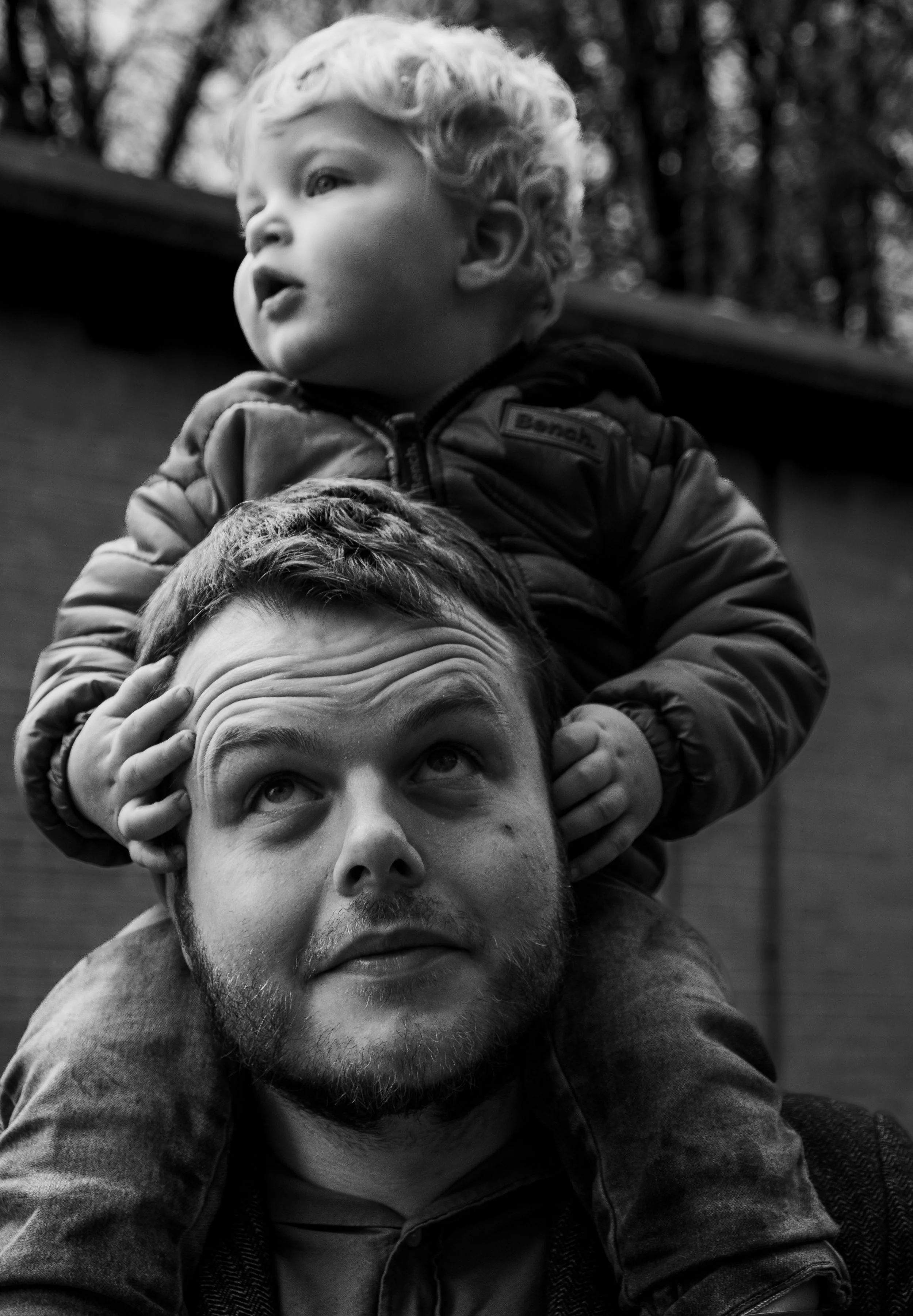 A man with facial hair lying on the ground, with a young child sitting on his shoulders, both looking upward in an outdoor setting with blurred trees in the background.