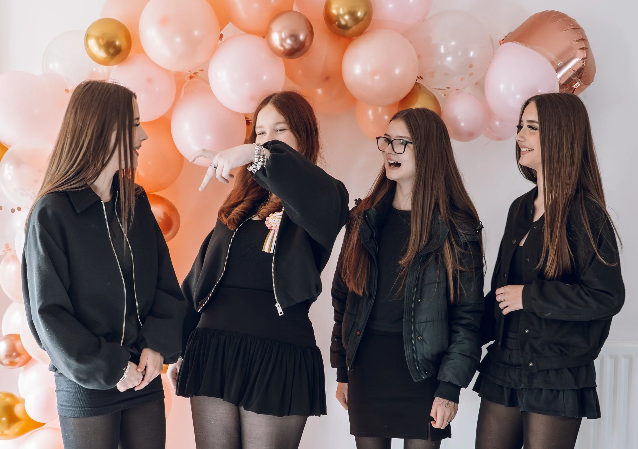 four teenage girls interact with each other showing happiness and comfort. they stand beneath a balloon arch and are pointed toward each other 