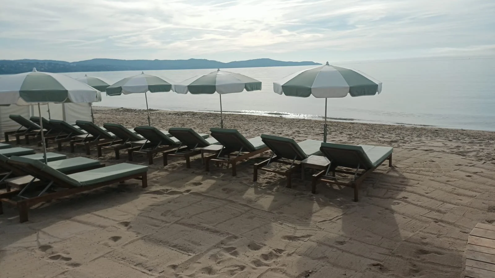 Chaises longues avec parasols sur une plage au bord de la mer, avec des montagnes en arrière-plan dans un ciel partiellement nuageux.