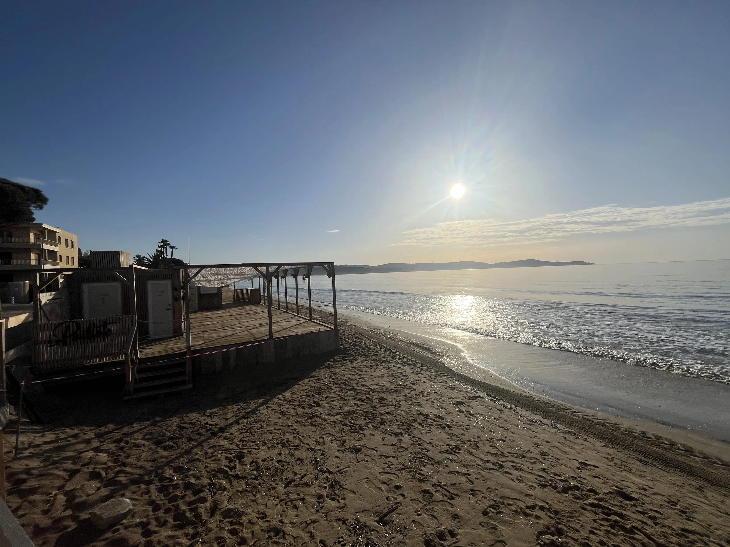 Une plage avec des bâtiments en arrière-plan, le soleil brillant dans le ciel, et des chaises longues et un parasol en bois sur le sable.
