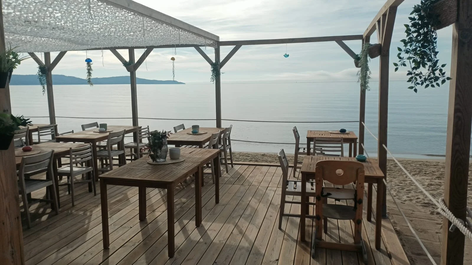 Terrasse en bois avec des tables et des chaises en face de la mer, décorée avec des plantes suspendues. La mer calme et un ciel nuageux en arrière-plan.