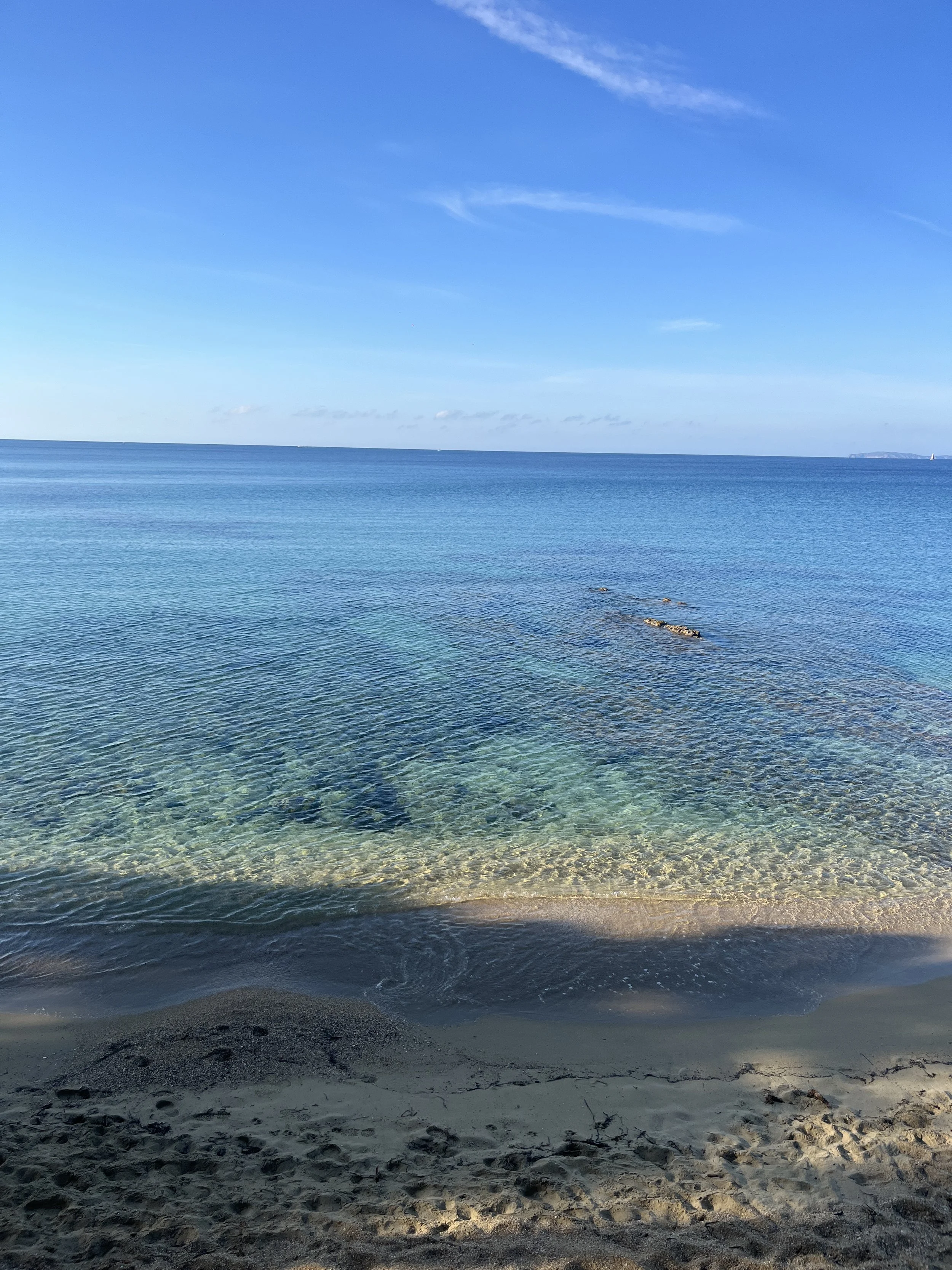 Une plage de sable avec des eaux calmes, sous un ciel bleu avec quelques nuages, vue lointaine de l'océan.