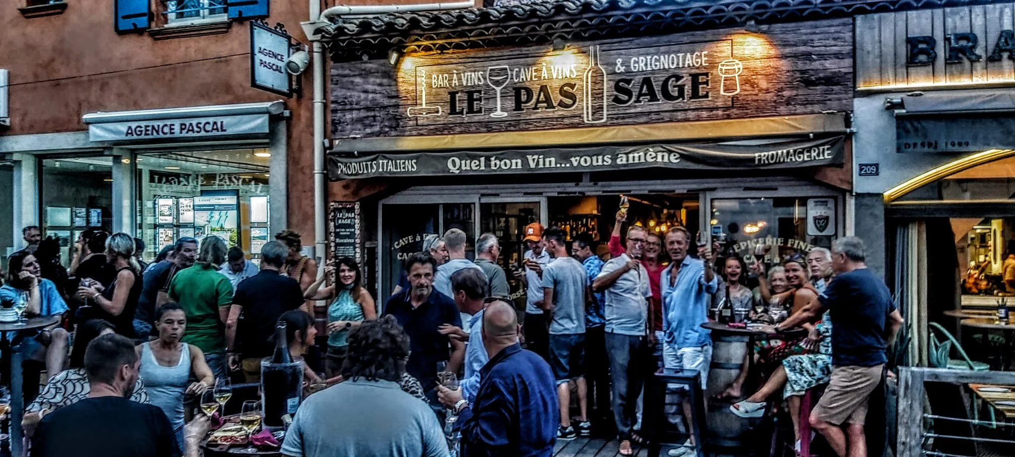 Une terrasse de bar animée avec des groupes de personnes dégustant des boissons, devant un bar avec un panneau indiquant 'Le Pas Sage', avec beaucoup de gens souriants et discutant.