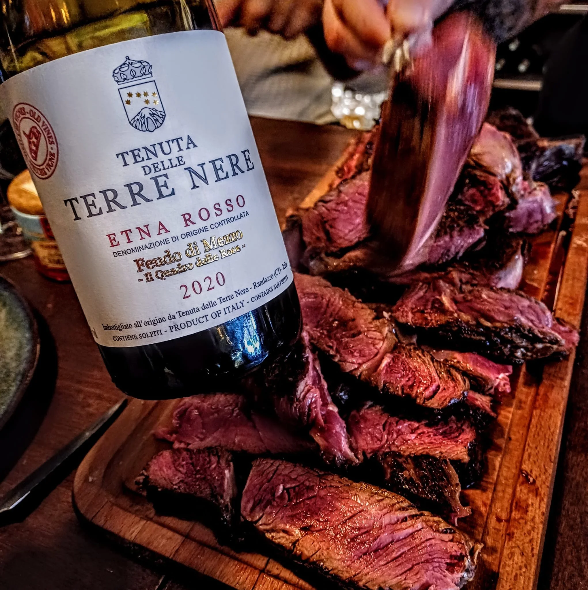 Une bouteille de vin rouge de la Tenuta delle Terre Nere posée sur une planche en bois, à côté d'une pièce de viande rouge cuite, dans un cadre chaleureux.