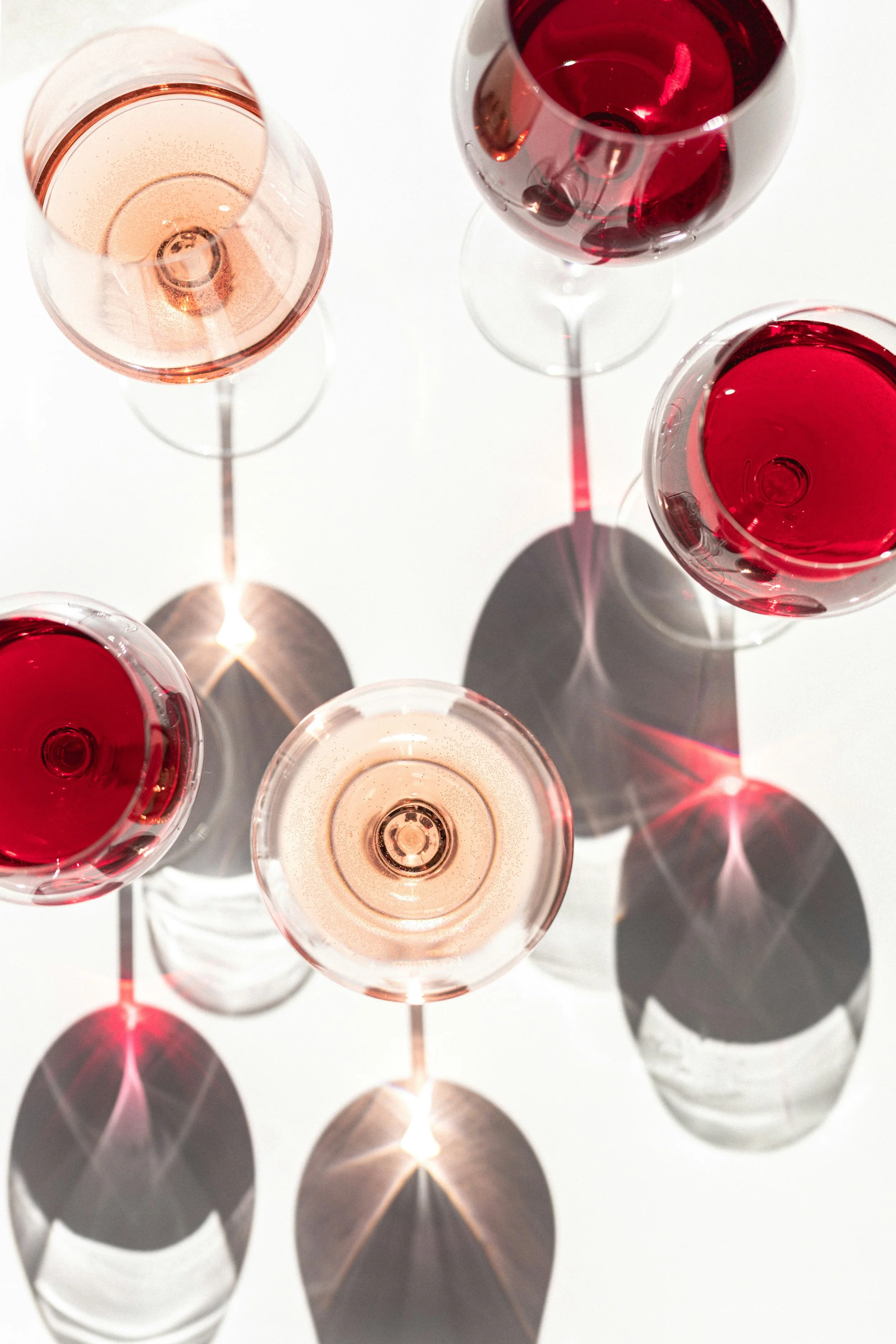 Coupes à vin contenant du vin rosé, rouge et blanc, avec leurs ombres projetées sur une surface blanche.
