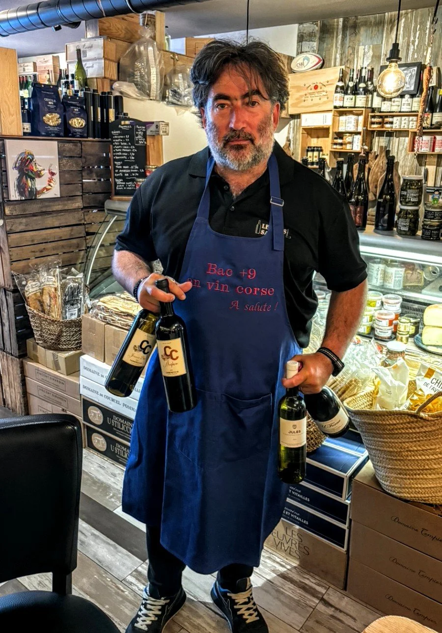 Homme dans une boutique, portant un tablier bleu avec une inscription en français, tenant des bouteilles de vin dans ses mains.