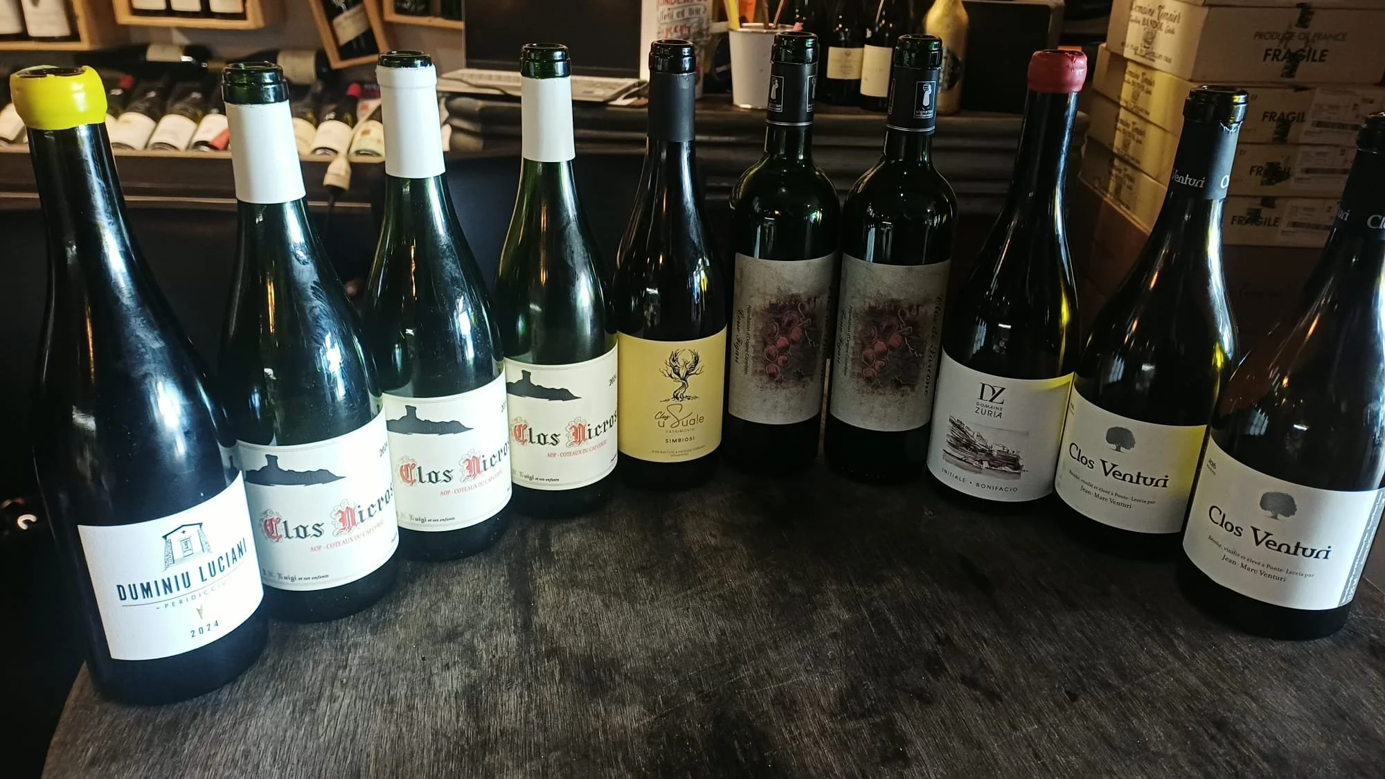 Une rangée de bouteilles de vin sur une surface en bois, avec divers étiquettes de vin visibles, dans une cave ou un magasin de vins.