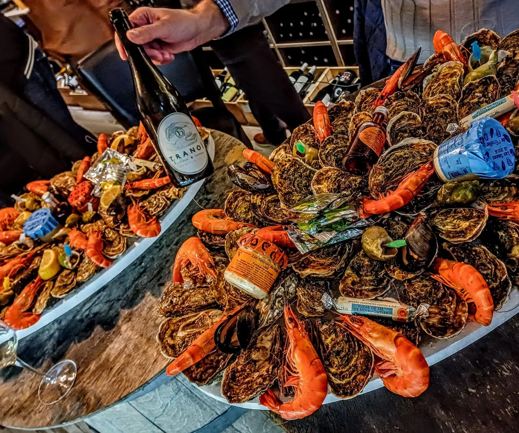 Plate d'huîtres garnies de crevettes, avec quelques bouteilles, sur un comptoir en bois, en salle de dégustation.