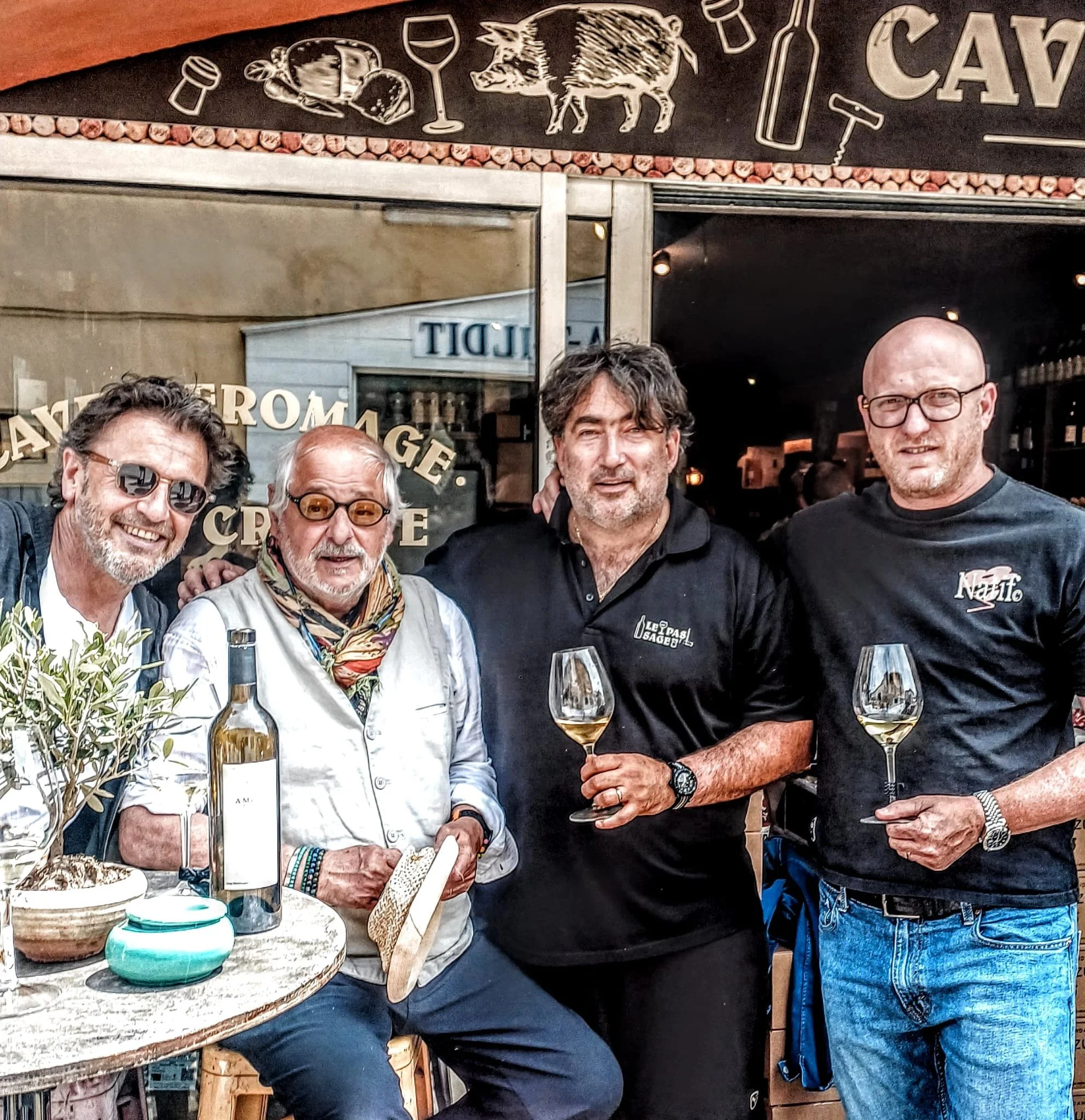 Groupe de cinq hommes posant pour une photo devant un restaurant ou bar, avec des verres de vin devant eux.