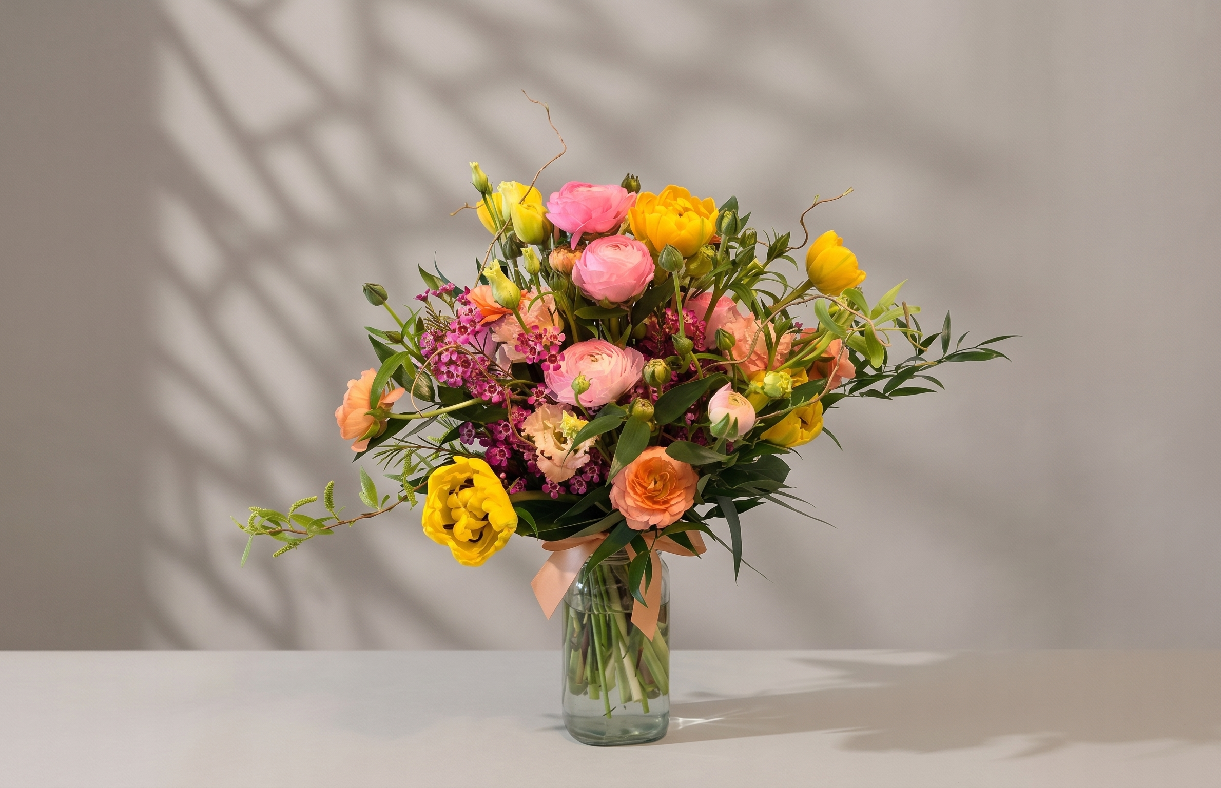 A bouquet of flowers in a glass vase on a cream background. The flowers are spring colours, yellow, orange and pinks and there is lots of greenery in it.
