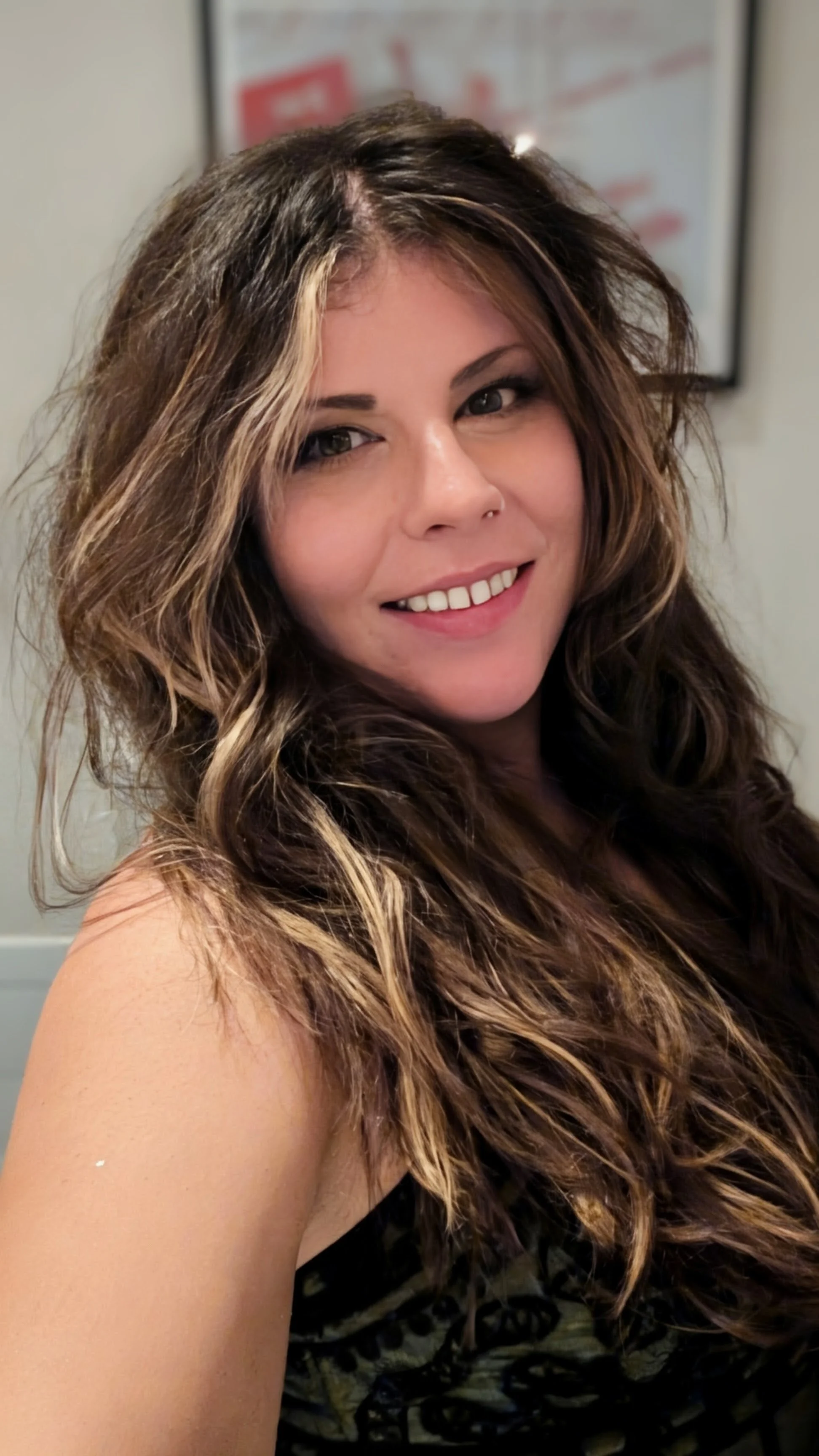 A woman with long, wavy hair and a smile, wearing a sleeveless top, standing indoors with a blurred background.