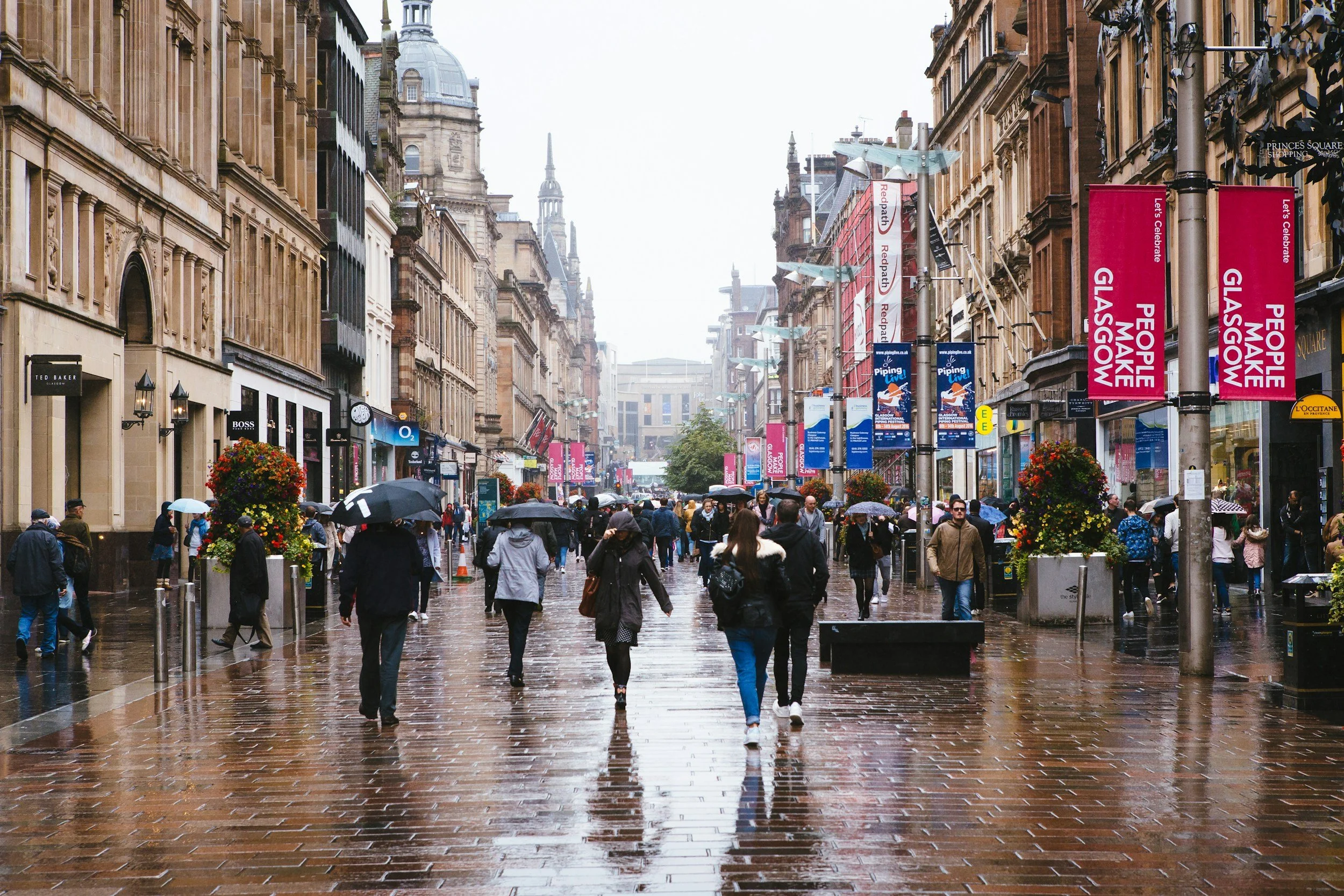 Crowd walking in Glasgow's Buchanan Street on a rainy day, with wet pavement, historic buildings, banners, and umbrellas.