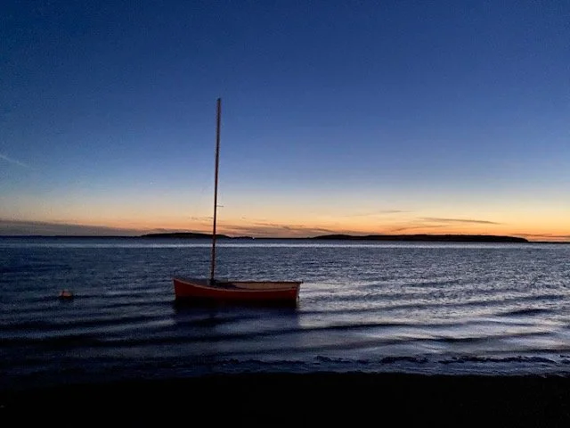 A sailboat floating on calm water during sunset with a clear sky and distant islands on the horizon.