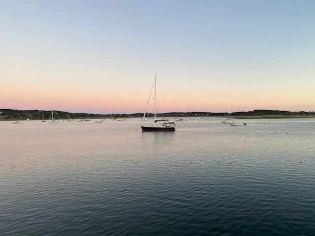 A calm body of water with a sailboat and several smaller boats at sunset, with low hills in the background.
