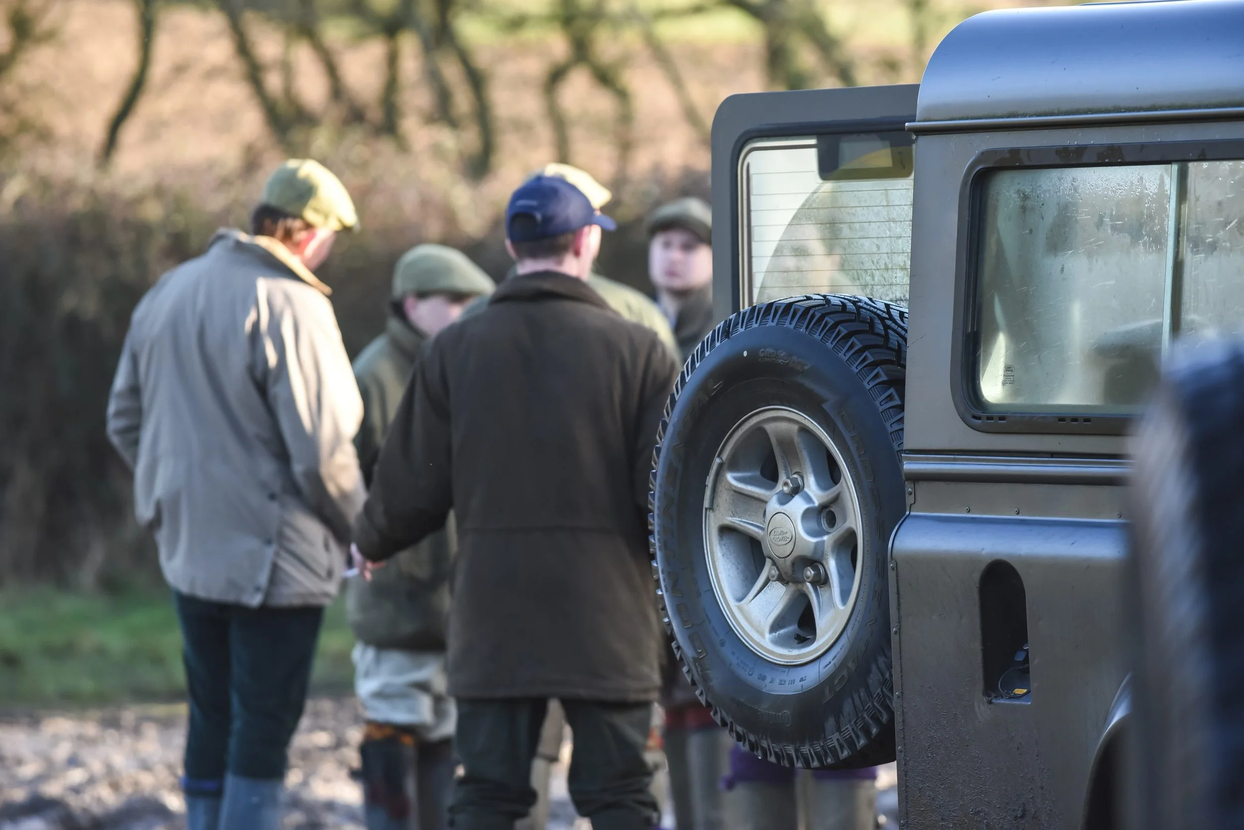 A group of guns standing in a circle outdoors near a vehicle with a spare tire, with trees in the background.