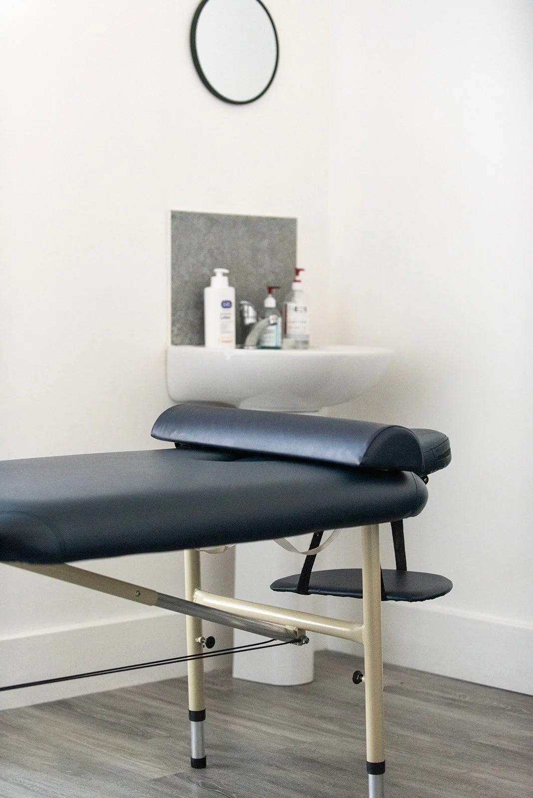 Physiotherapy treatment room with navy treatment table, small white sink with soap and sanitizer bottles, and a round mirror on a white wall.