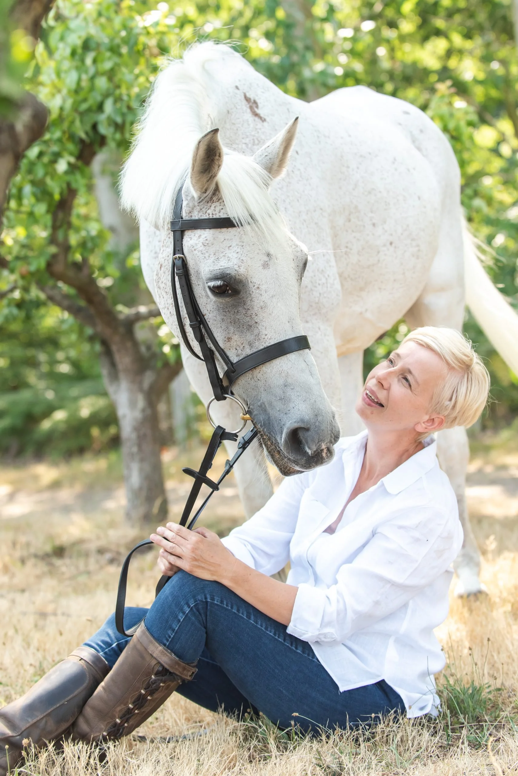 A woman with short blonde hair sitting on the ground, smiling and looking at a white horse with gray speckles. The woman is wearing a white shirt, jeans, and brown riding boots. The horse has a black halter and is nuzzling the woman. They are outdoor