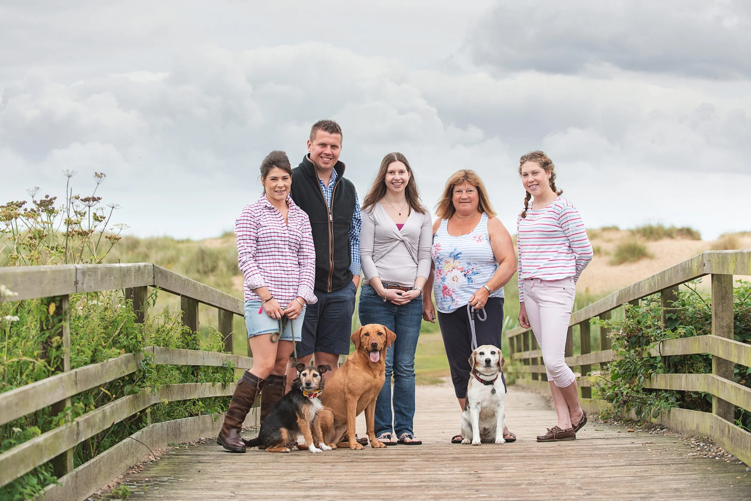 A group of five people with six dogs standing on a wooden bridge outdoors, with green plants on either side and cloudy sky overhead.