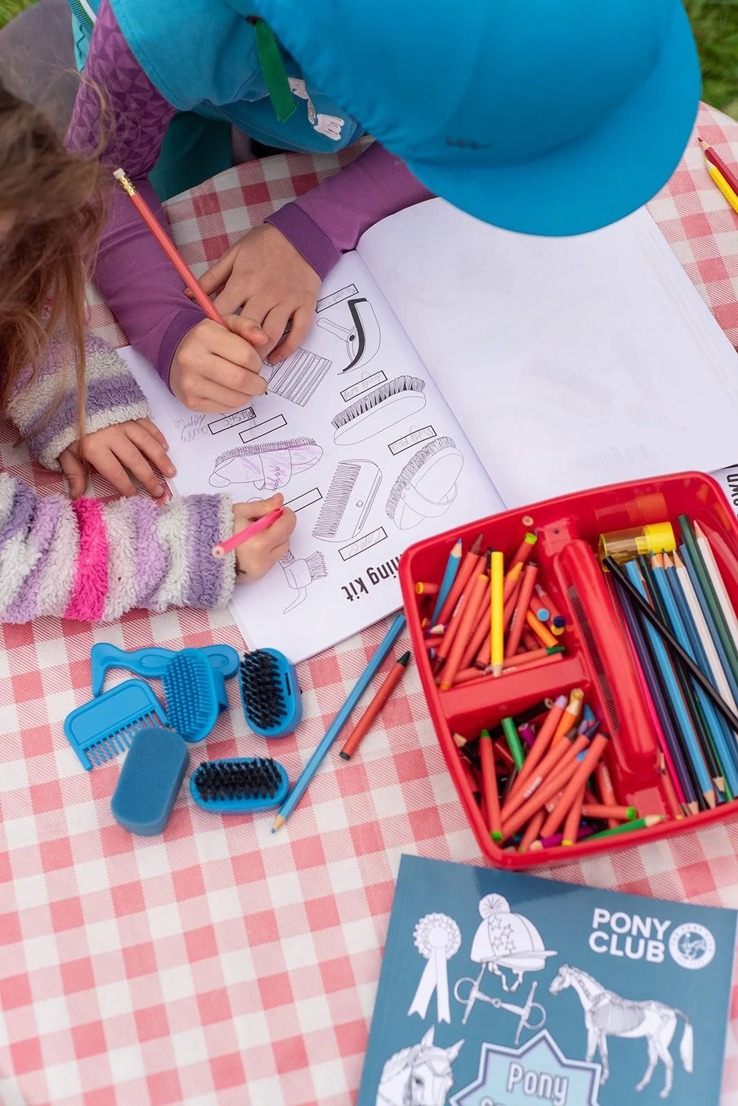 Two children coloring a grooming kit illustration in an open coloring book on a checkered pink and white tablecloth, surrounded by colored pencils and grooming tools.