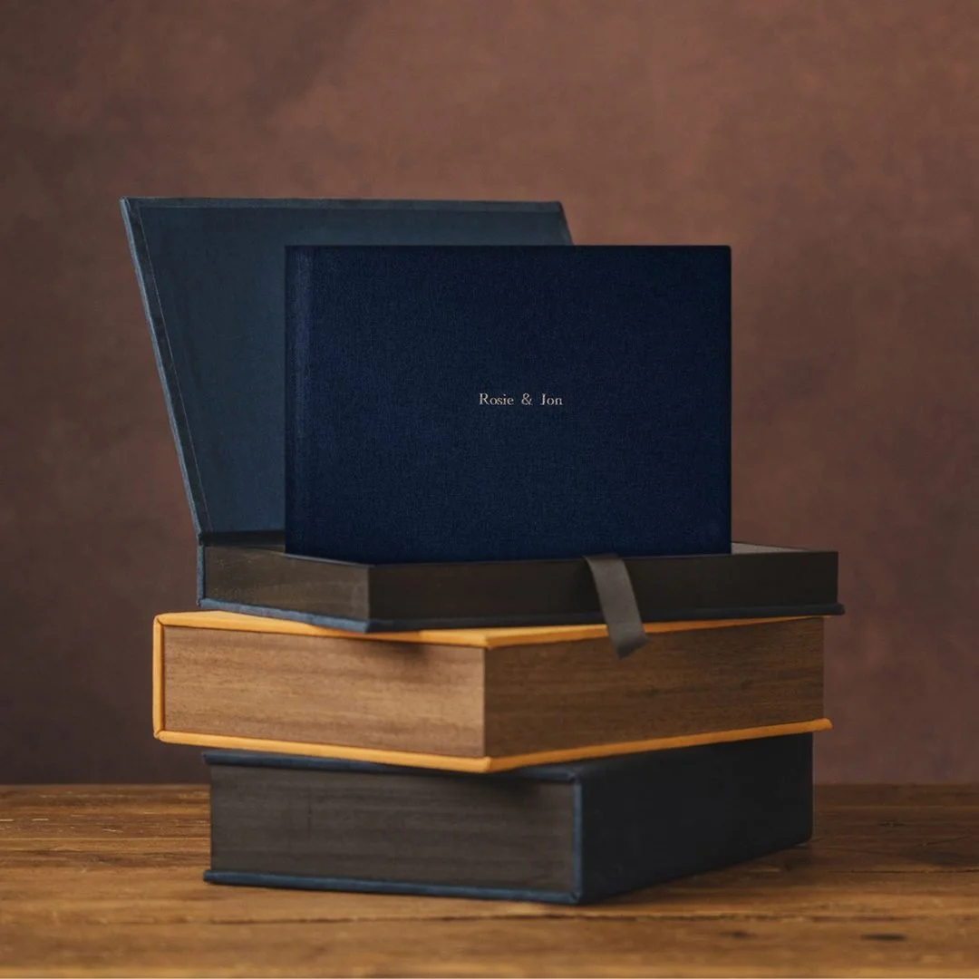 Stack of three photo album display boxes on a wooden surface, with the top one open and a blue box beneath it. The second book has a light brown cover. The background is dark brown.
