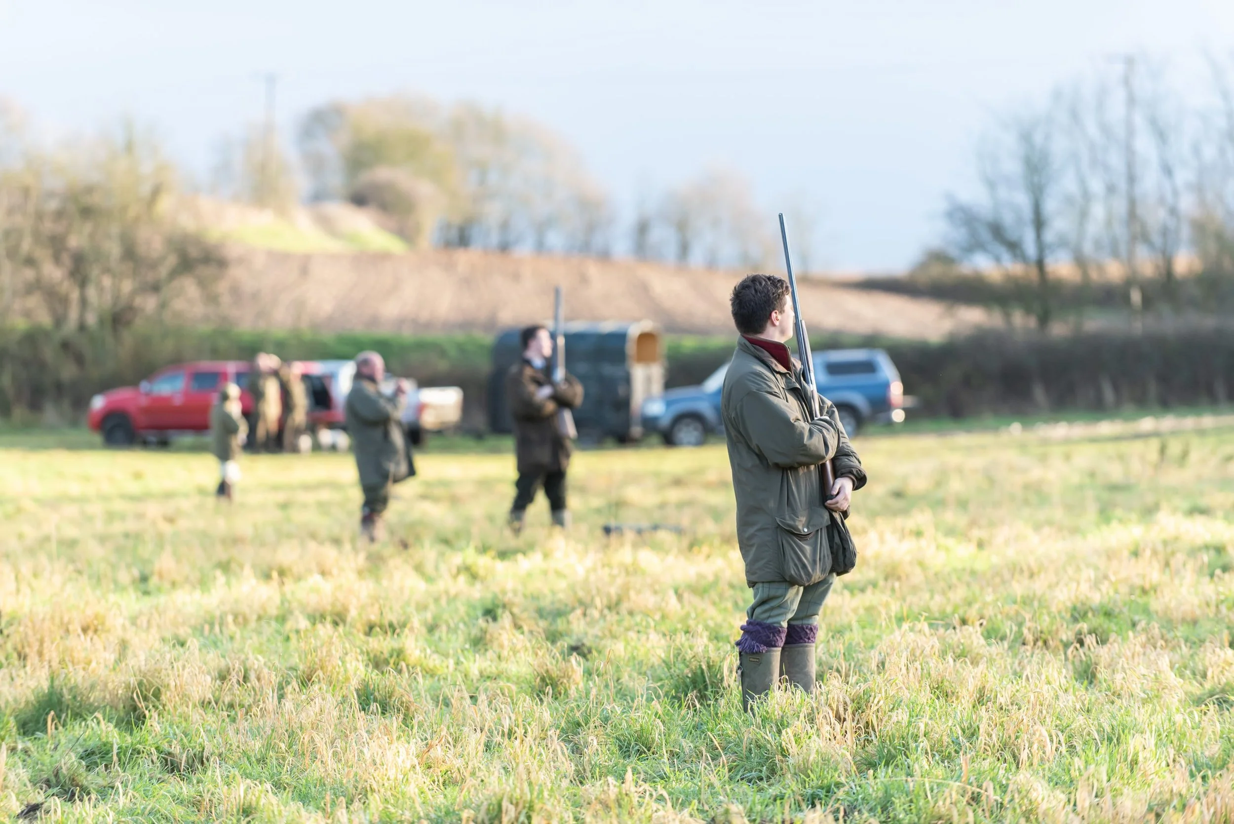 A young man standing in a grassy field holding a shotgun, with several other people in the background, some also holding firearms, near parked vehicles on a cloudy day.