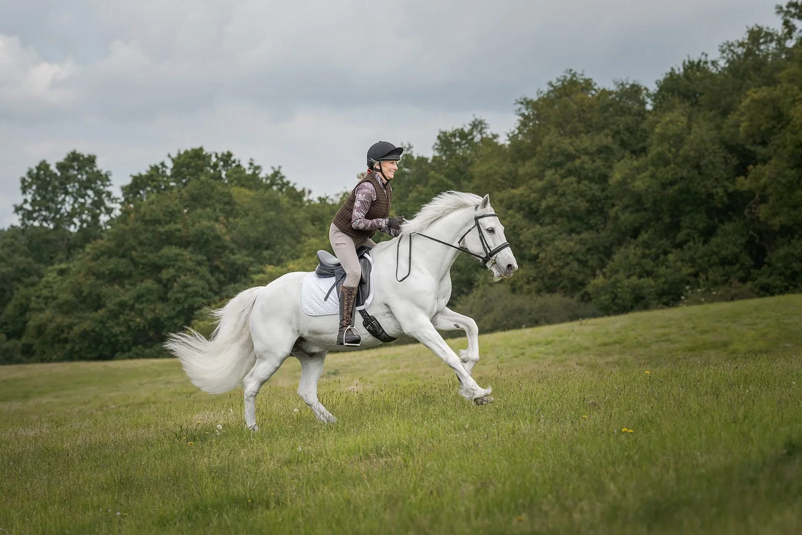 Woman riding a white horse across a grassy field on a cloudy day.